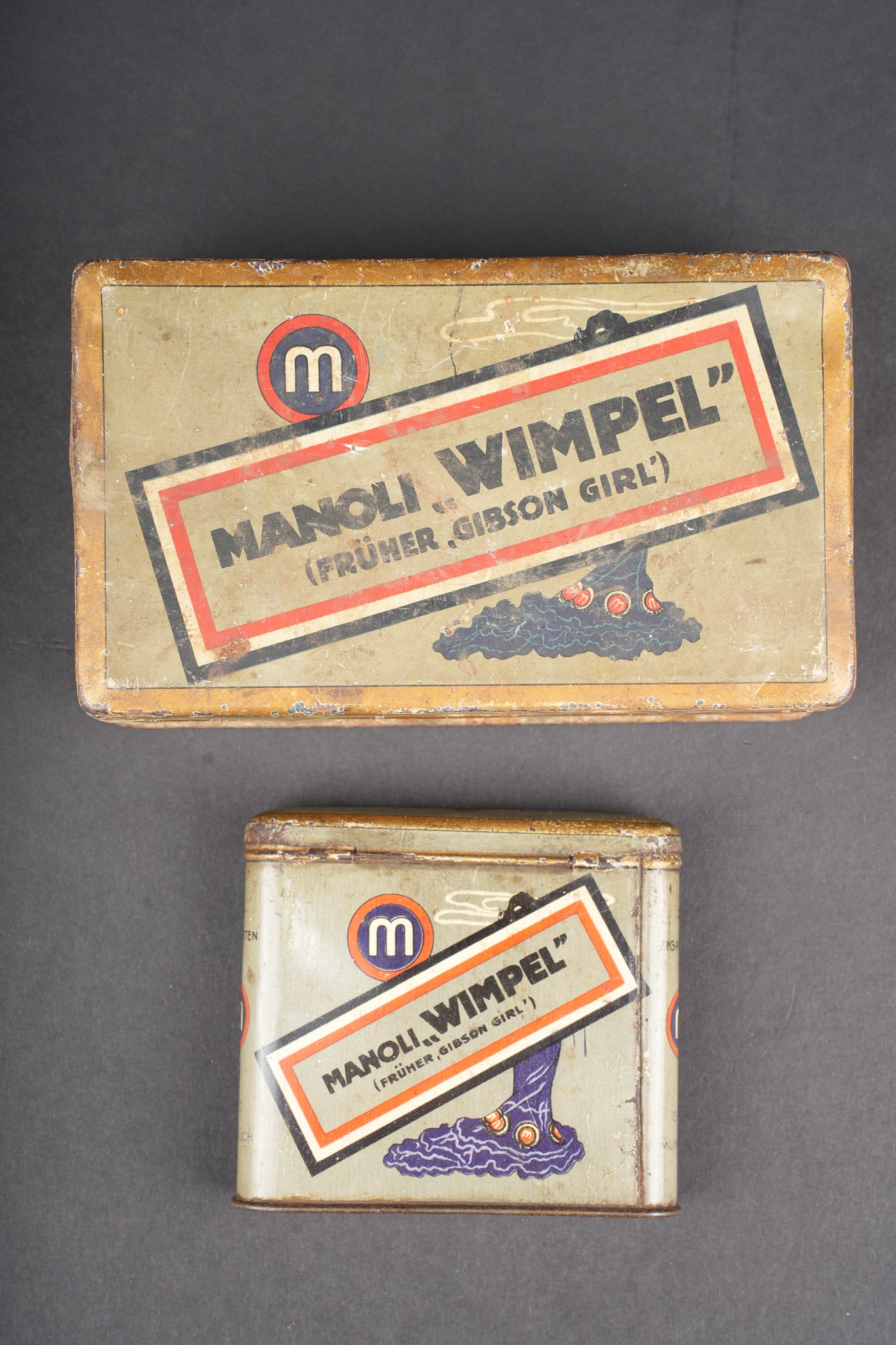 Lot de 2 boites de cigarettes allemandes Manoli Wimpel. Lot of two German Manoli Wimpel cigarette: Lot de 2 boites de cigarettes allemandes Manoli Wimpel. Toutes deux fond feldgrau et motifs aux couleurs nationales. Initialement marque Gibson Girl, juge trop anglais et change durant la guerre en
