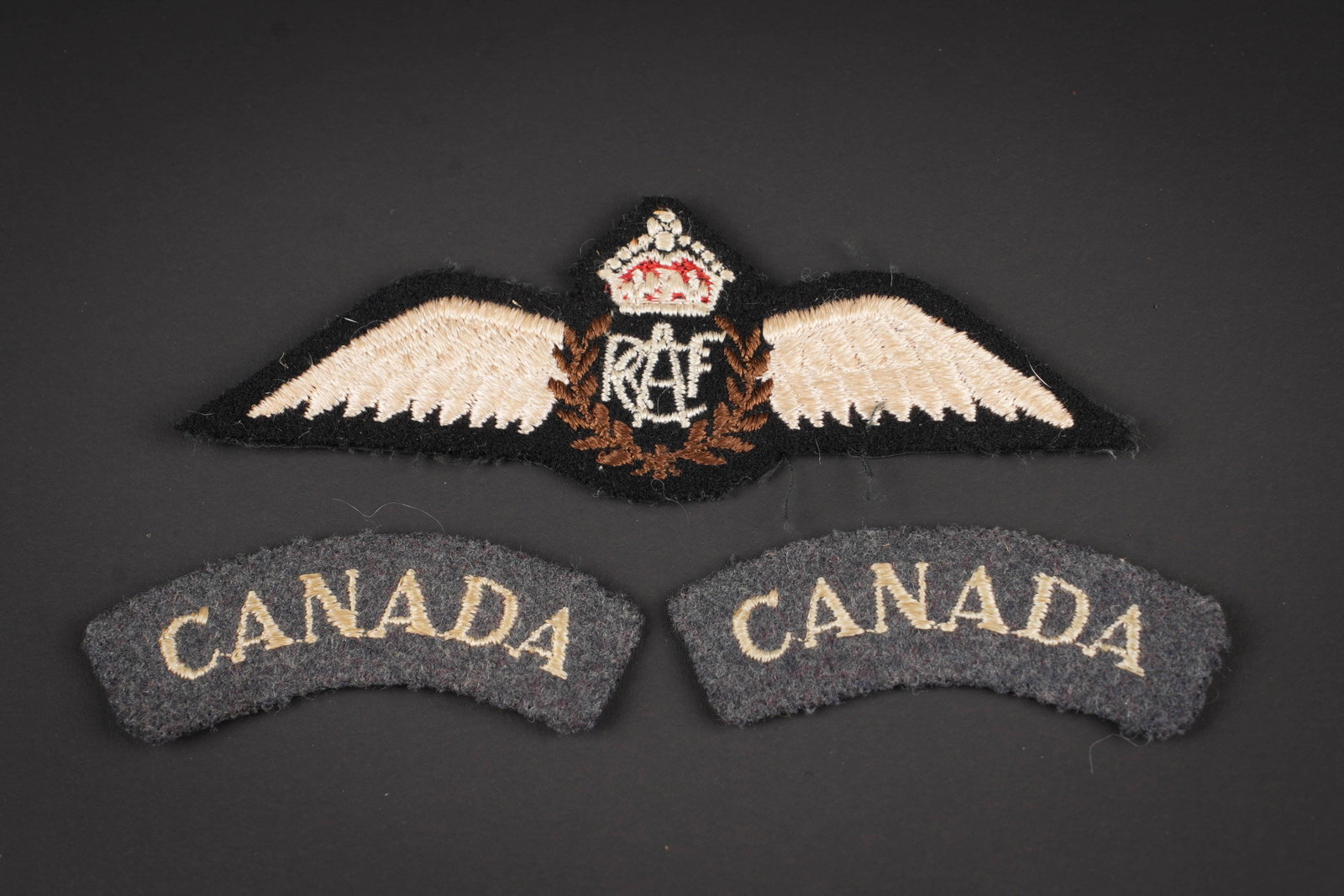Insignes RCAF. RCAF insignias.: Insignes RCAF. Comprenant deux titles en drap bleu, broderie marquee Canada. Brevet de pilote de la RCAF, dos de la piece double de tissu coton noir. Modele de fabrication a plat. A noter une legere