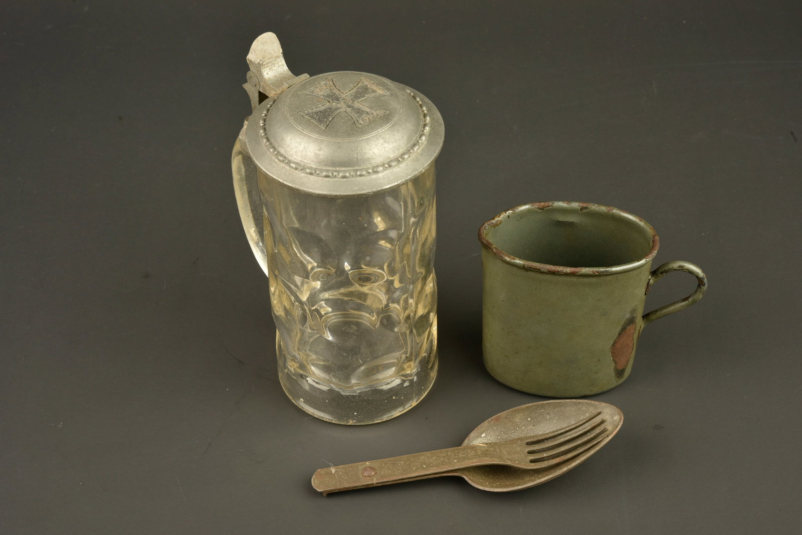 Lot allemand chope, quart et couverts. German lot including stein, drinking cup and cutlery: Lot allemand chope, quart et couverts. Une chope en verre epais avec couvercle en etain a motif de croix de fer. La piece de pouce a ete reparee. Un quart emaille vert avec logo fabricant 2 cimeterres