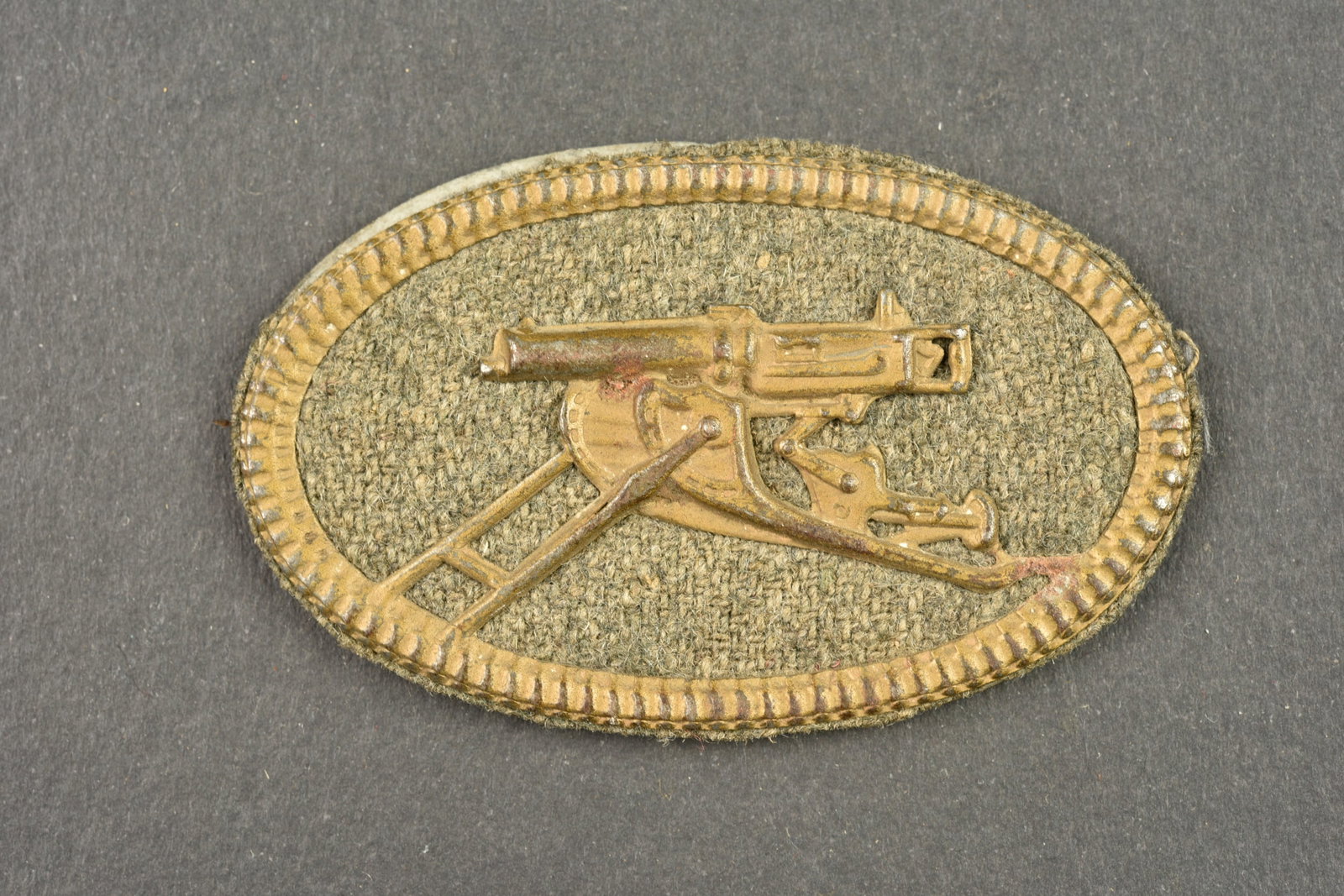 Insigne de mitrailleur allemand. German machine gunner badge: Insigne de mitrailleur allemand. Modele en fer peint feldgrau sur fond de drap feldgrau. Contre plaque marquee C.E. Junker Berlin. Une des patte arriere est cassee mais l insigne tient fermement. Bel 