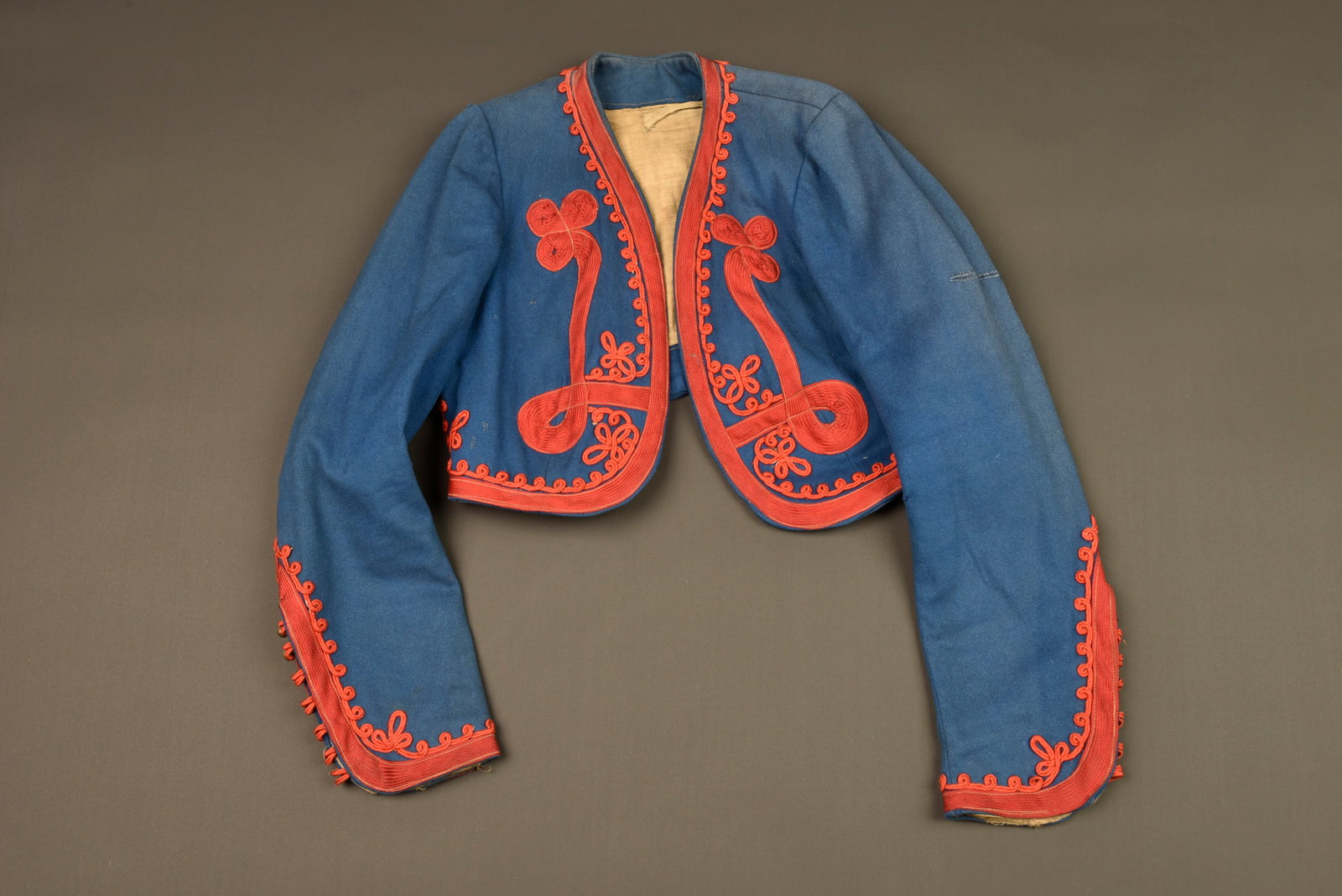 Bolero de zouave enfant. Child’s Zouave bolero jacket.: Bolero de zouave enfant. Confectionne en laine bleu de ciel, tresses et decors au fil de coton garance. Doublure en coton ecru portant le tampon du costumier Martin. Une reparation au niveau du coude 