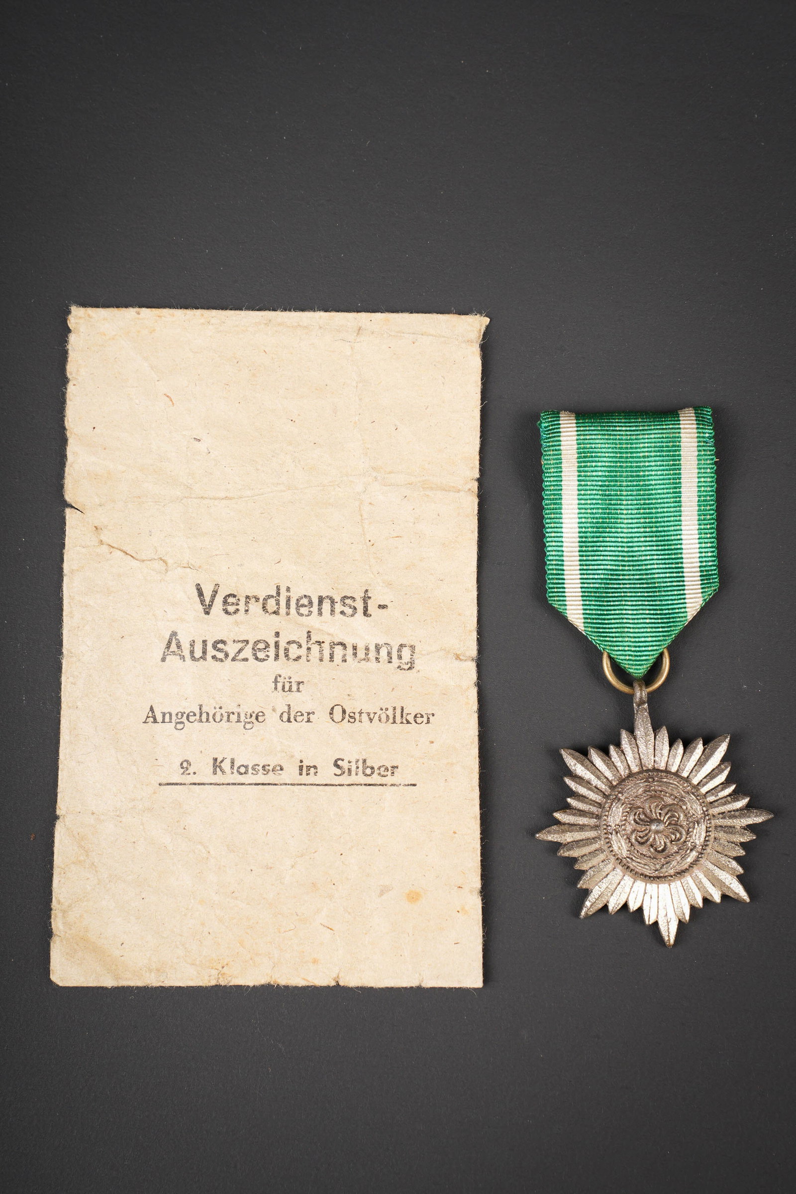 Ostvolker Medal. German medals.: Ostvolker Medal. Medaille en metal, couleur argent a 50%. Ruban complet. Attache complete. Sachet present, mais fortement abime. Marque Verdienst Auszeichnung fur Angehorige Der Ostvolker 2 Klasse in