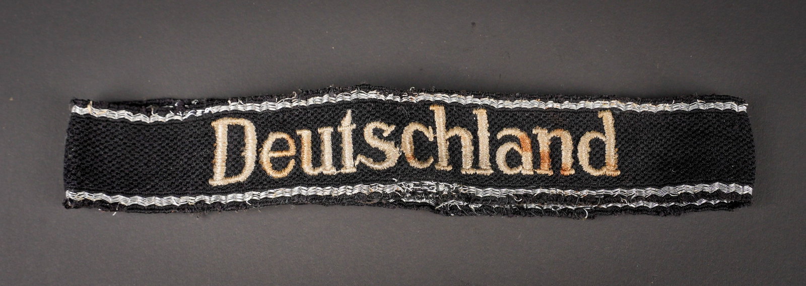 Bande de bras Deutschland. Deutschland cuff title.: Bande de bras Deutschland. En tissu noir disposant d un double lisere argent. Celui-ci est partiellement effiloche. Marquage brode Deutschland. Bande de bras cousue et visiblement demonte d une