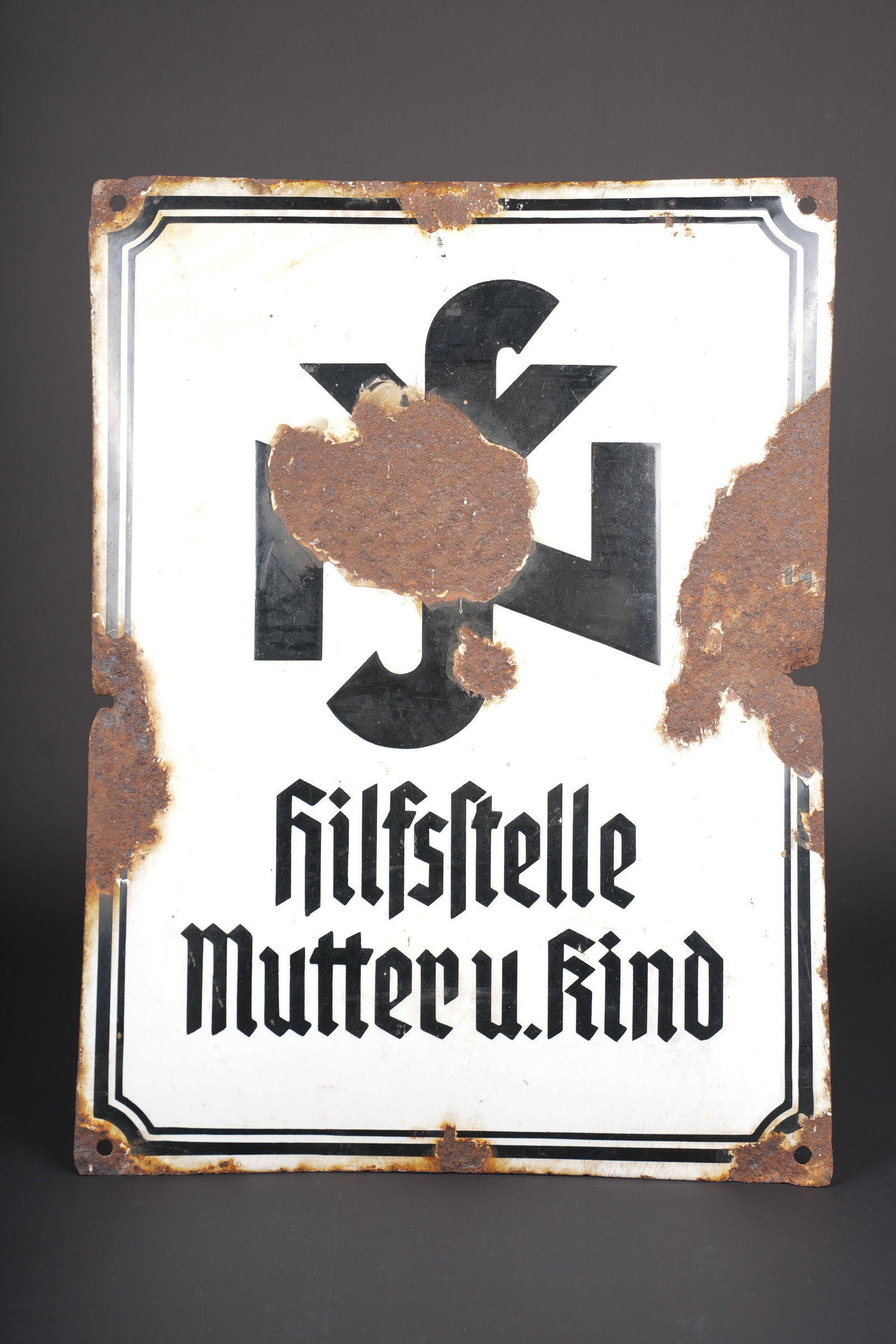 Plaque emaillee Hilfstelle. Hilfsstelle enamel sign.: Plaque emaillee Hilfstelle. En metal emaille bombe. Fond de couleur blanc, marquage noir Hilfstelle Mutter U Kind. Nombreux manques sur les bordures. Eclats important dans l email. Quatre points de