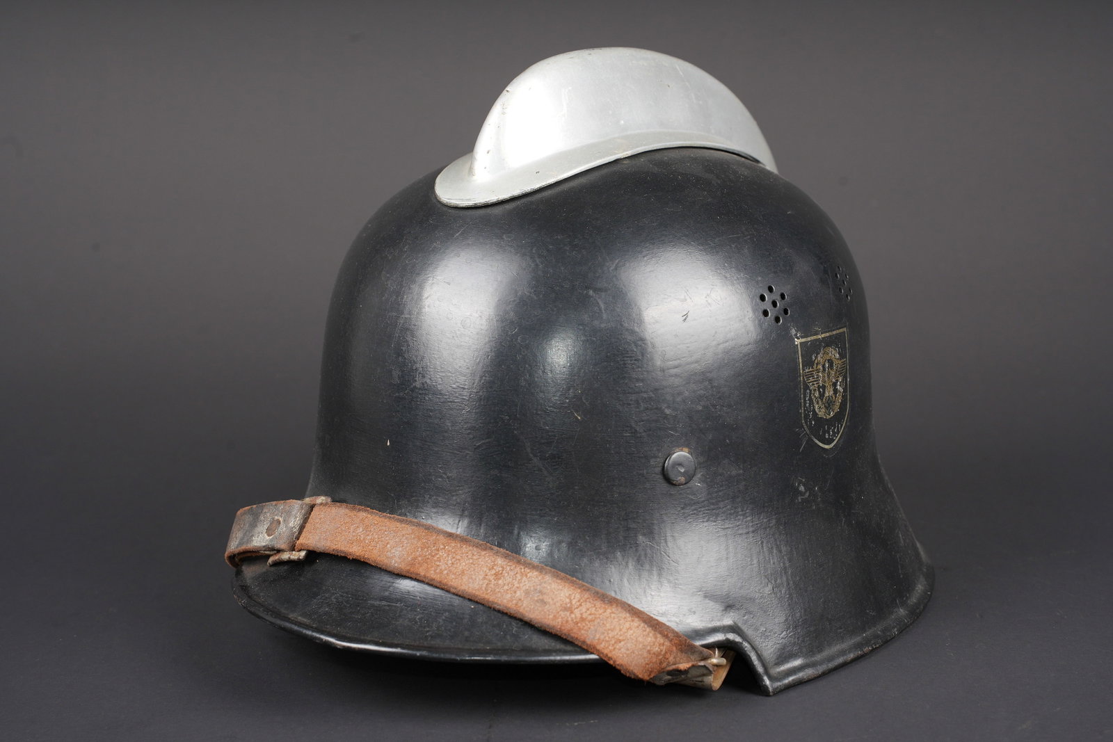 Casque Feuerschutzpolizei. Feuerschutzpolizei helmet.: Casque Feuerschutzpolizei. Modele 34. Peinture noire a 80%. Cimier en aluminium couleur chrome. Les insignes sont des decalcomanies, presents a 70 et 80%. Jugulaire en cuir marron. La nuquiere