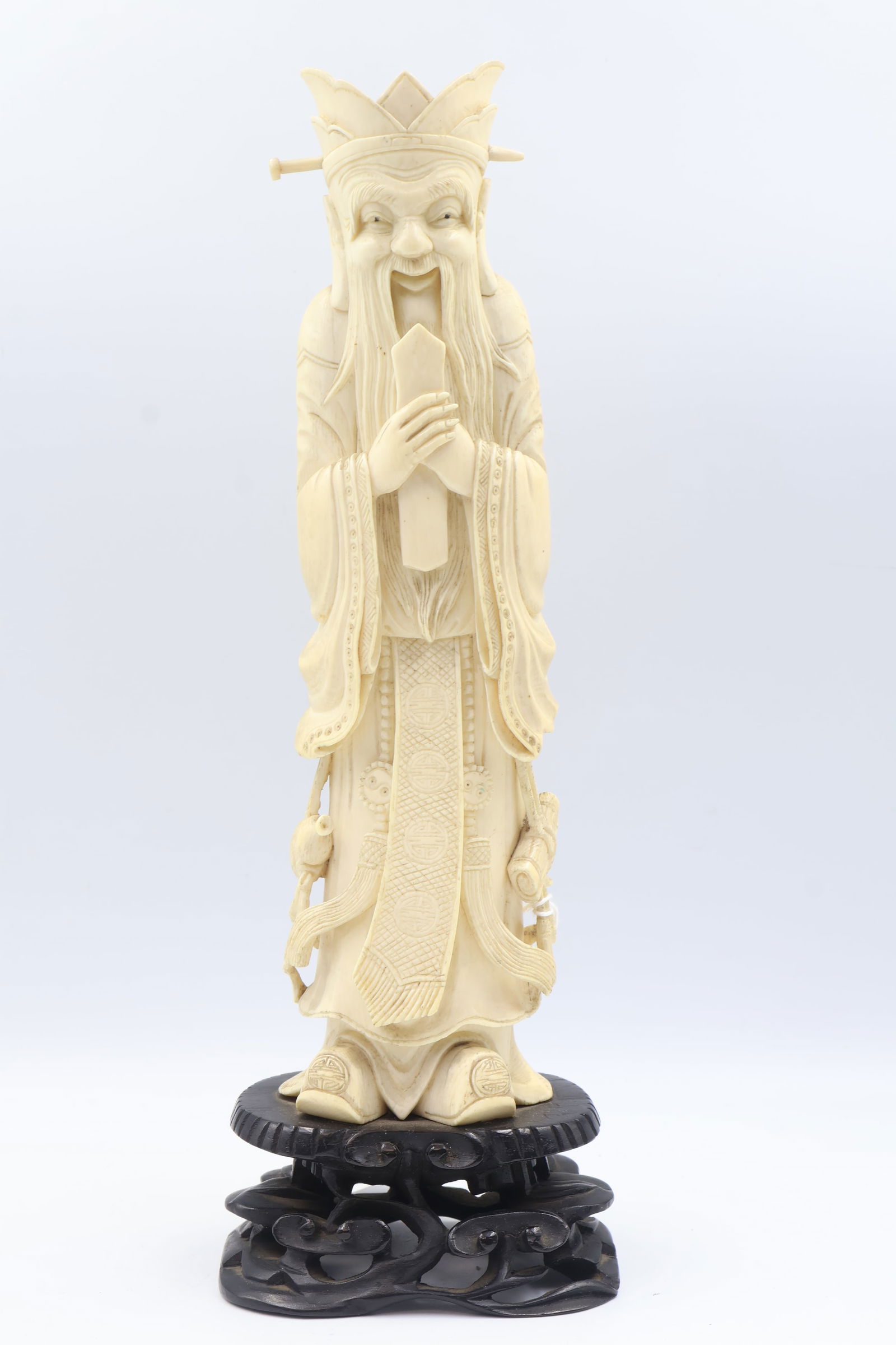 An antique hand carved Chinese bone figural study.: The standing crowned sage or scholar is in traditional draped attire bearing a hanging vessel, scroll and a sash featuring happiness symbols. The whole is mounted to a carved & pierced hardwood stand.