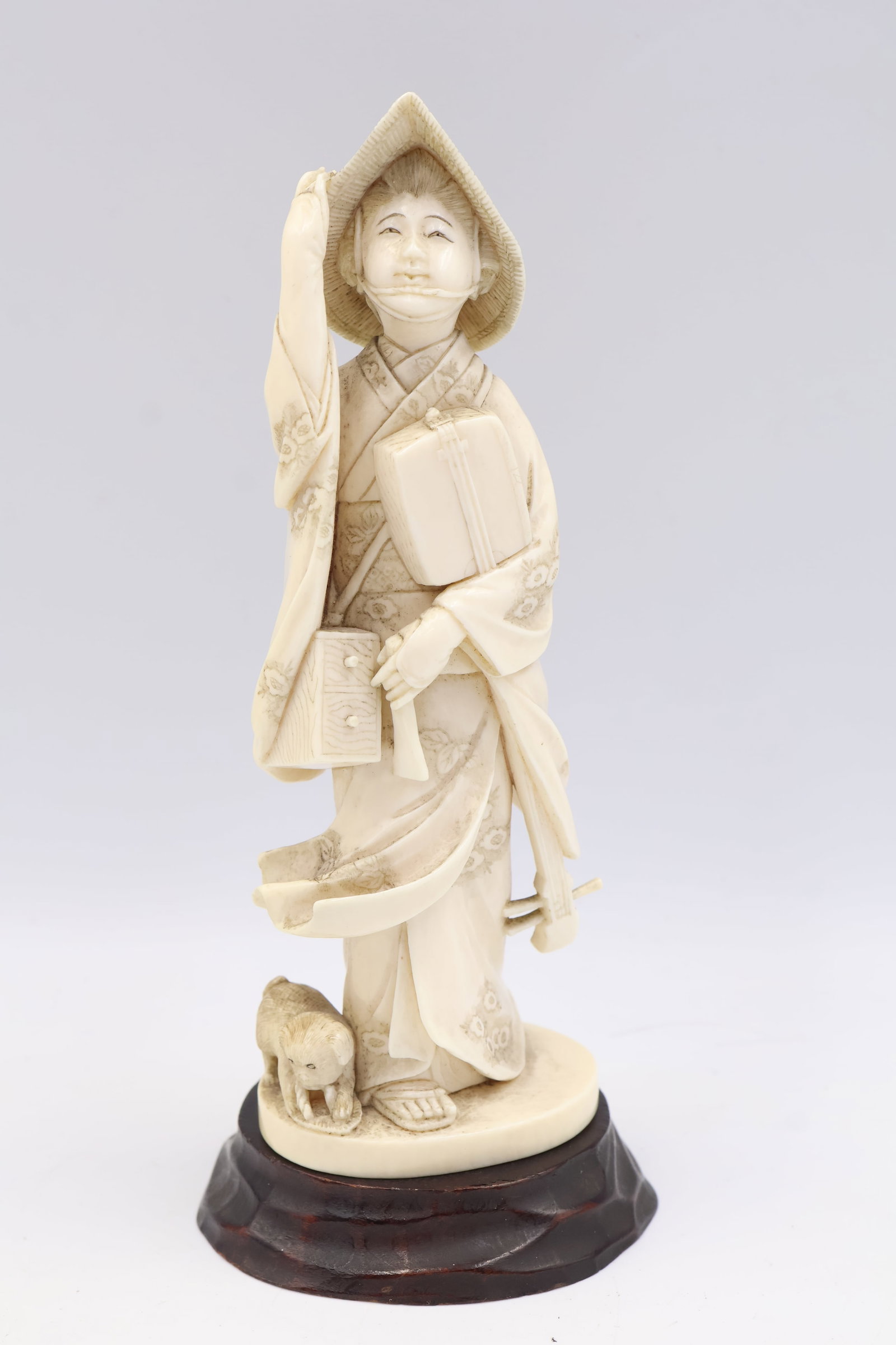 Hand Carved JapaneseIvory Okimono: The windswept standing figure (possibly a traveller or merchant) holding her hat features boxes, tools and a Shamisen. Red lacquer seal signature underside, Meiji period. Dimensions: 6” height