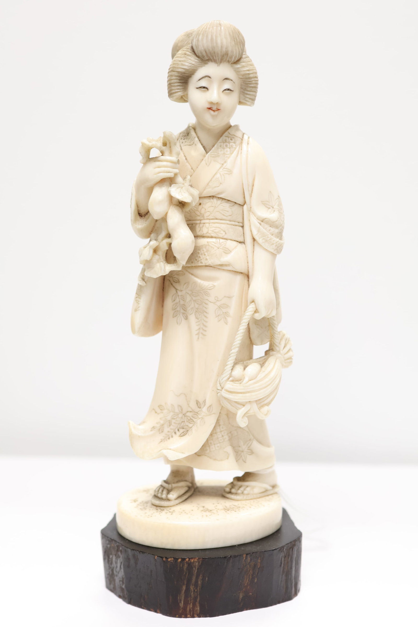 A Signed, Hand Carved Japanese Okimono: This beautifully executed carving depicts a Bijin (beautiful woman or Geisha) dressed in traditional kimono holding a basket of produce. Red lacquer seal signature underside, Meiji period.