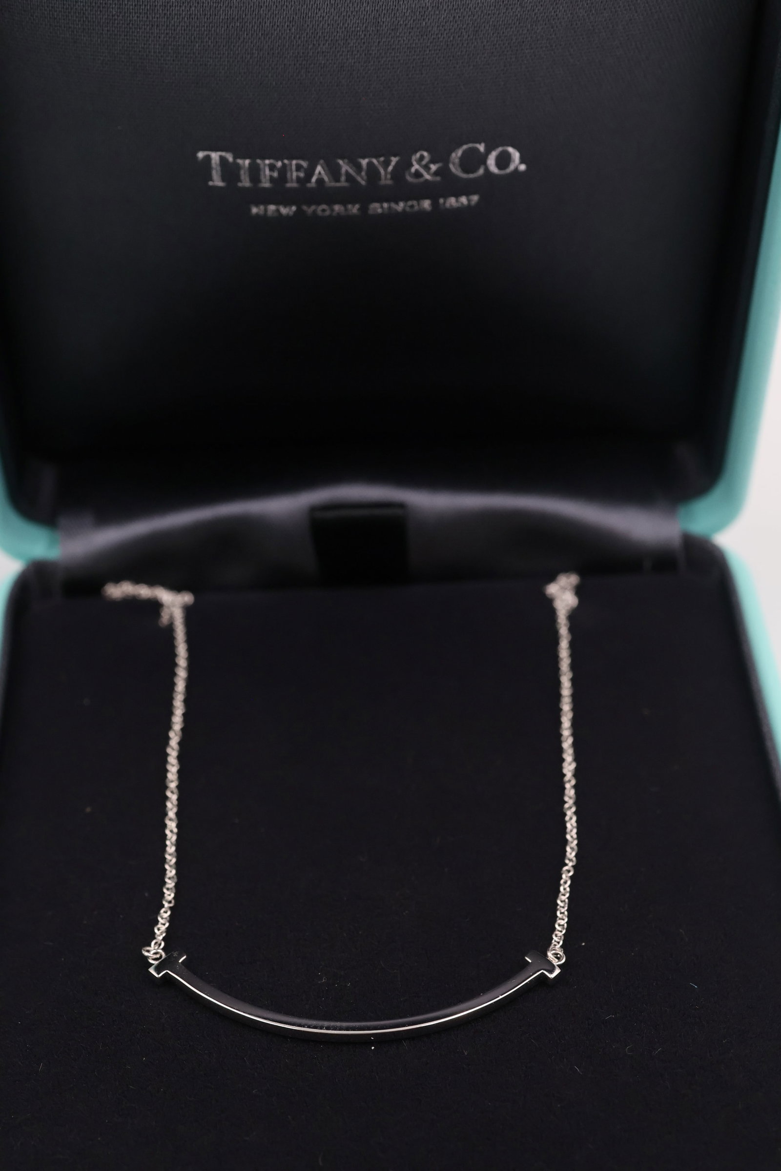 Tiffany & Co 18k White Gold Smile Necklace: Tiffany & Co. Smile necklace crafted in 18K white gold, featuring its distinctive curved silhouette. Comes with boxes, bag and charm. Comes with original box. Weight 3.08 grams.