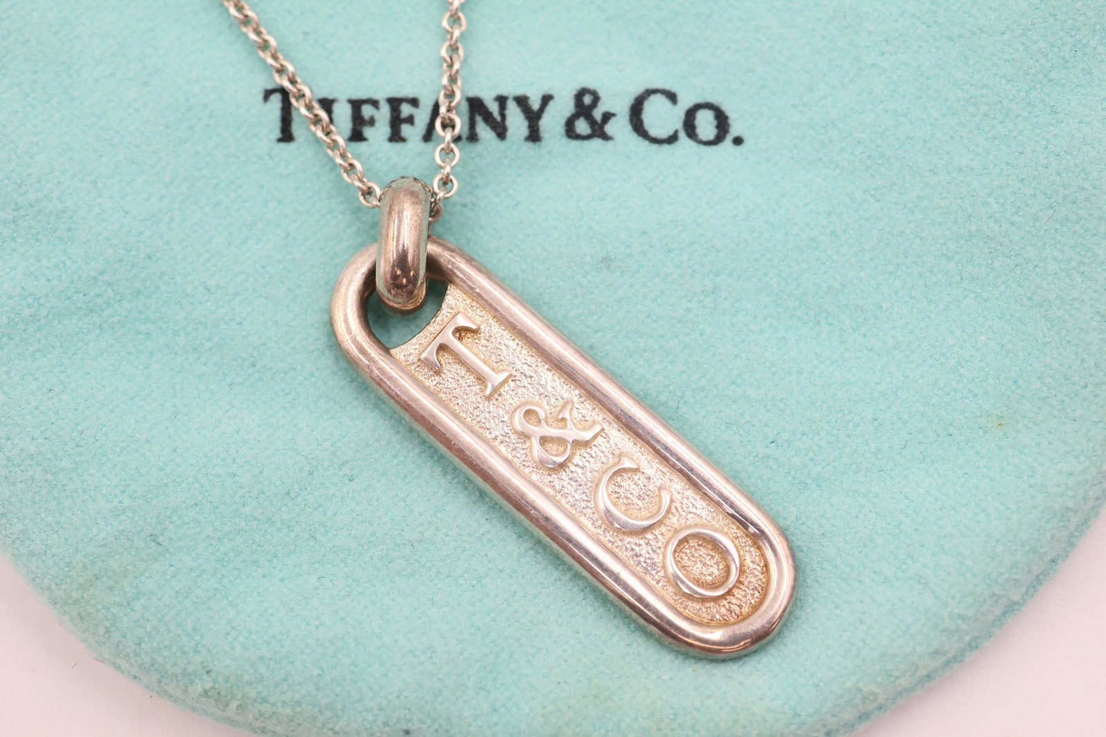Tiffany & Co 925 Oval Logo Tag Pendant & Chain: Tiffany & Co. necklace featuring a signature oval logo tag pendant on a sterling silver chain. Total weight: 9.7 grams. Accompanied by a Tiffany & Co. pouch.