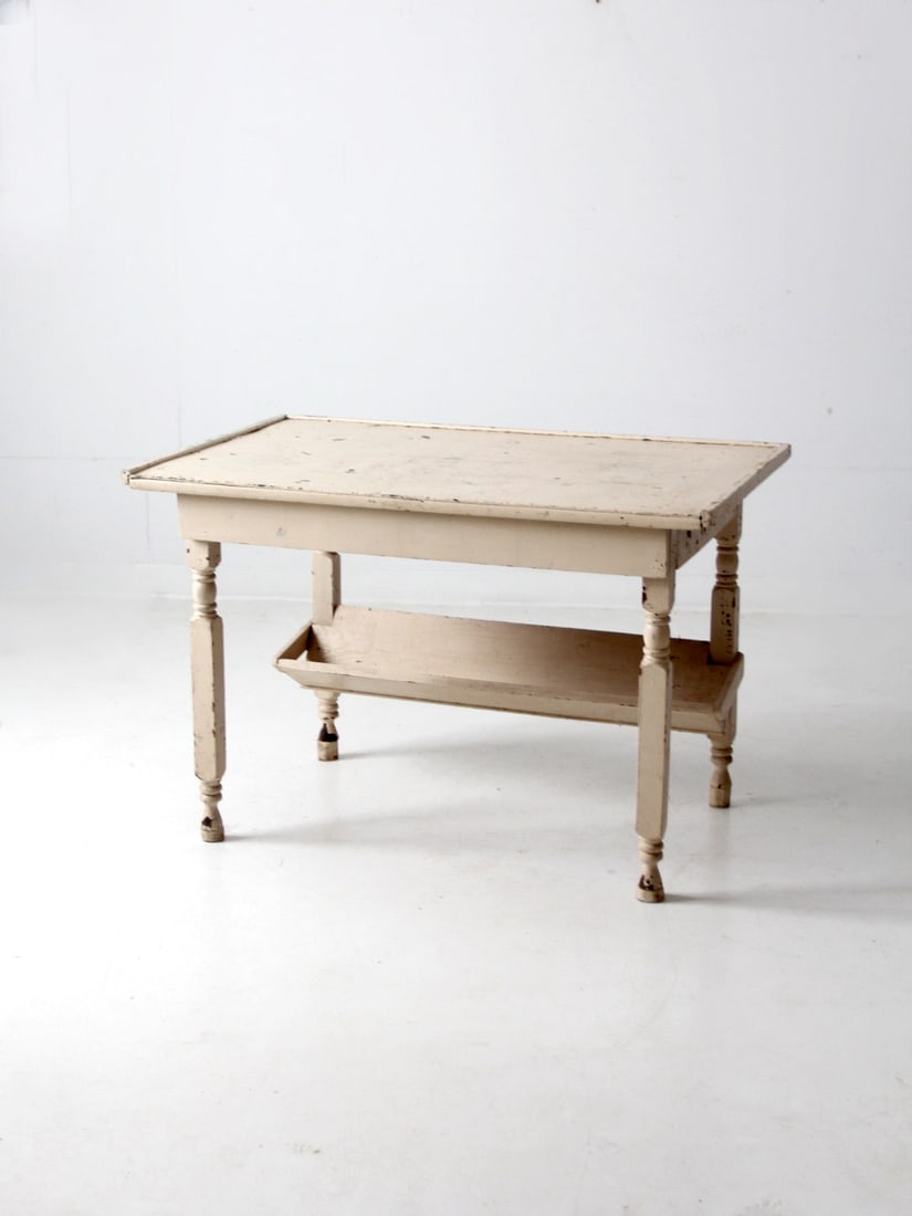 Antique Wooden Table with Trough Shelf and Creamy White Finish: Antique Wooden Table with Trough Shelf and Creamy White Finish This antique wooden table boasts a beautifully weathered creamy white finish and elegant turned legs. The tabletop features a lipped edge