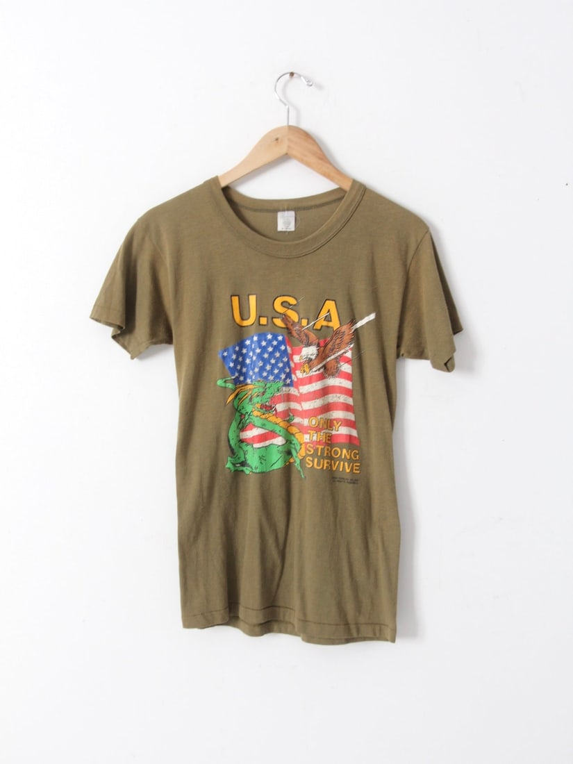 1987 Vintage USA Graphic Tee Eagle Dragon Design: 1987 Vintage USA Graphic Tee Eagle Dragon Design Discover a piece of the 1980s with this Vintage USA Graphic Tee. This ultra-soft olive green t-shirt features a striking design of an eagle and a drago