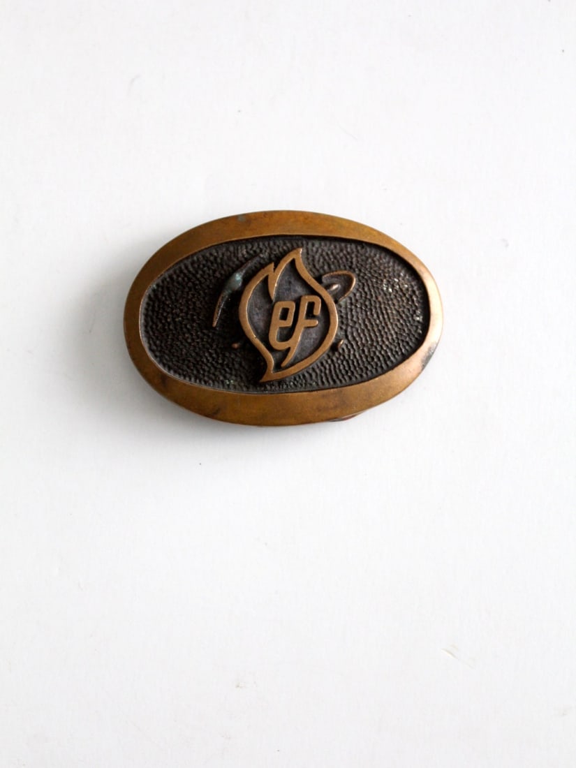 Vintage Brass Oval Plate Buckle with EF Flame Logo 3.5 in: Vintage Brass Oval Plate Buckle with EF Flame Logo 3.5 in Introducing a vintage brass buckle, showcasing an elegant oval plate style adorned with an EF flame logo across its face. This unique accessor