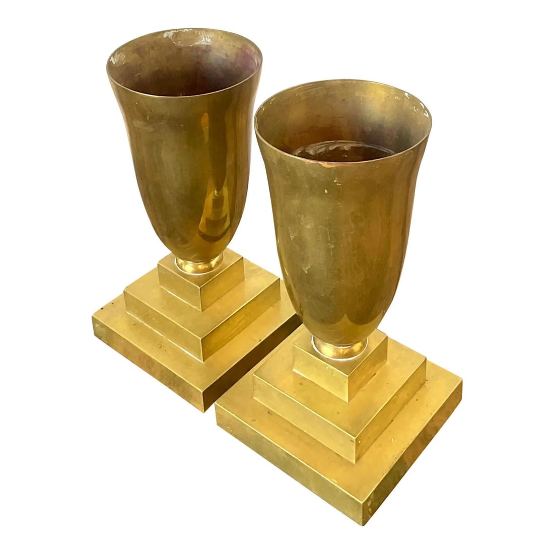 Pair of Brass Pedestal Urns: Pair of Brass Pedestal Urns This pair of vintage brass pedestal urns showcases classic Hollywood Regency style with Art Deco-inspired geometric design. Each urn features a stepped square base and a fl