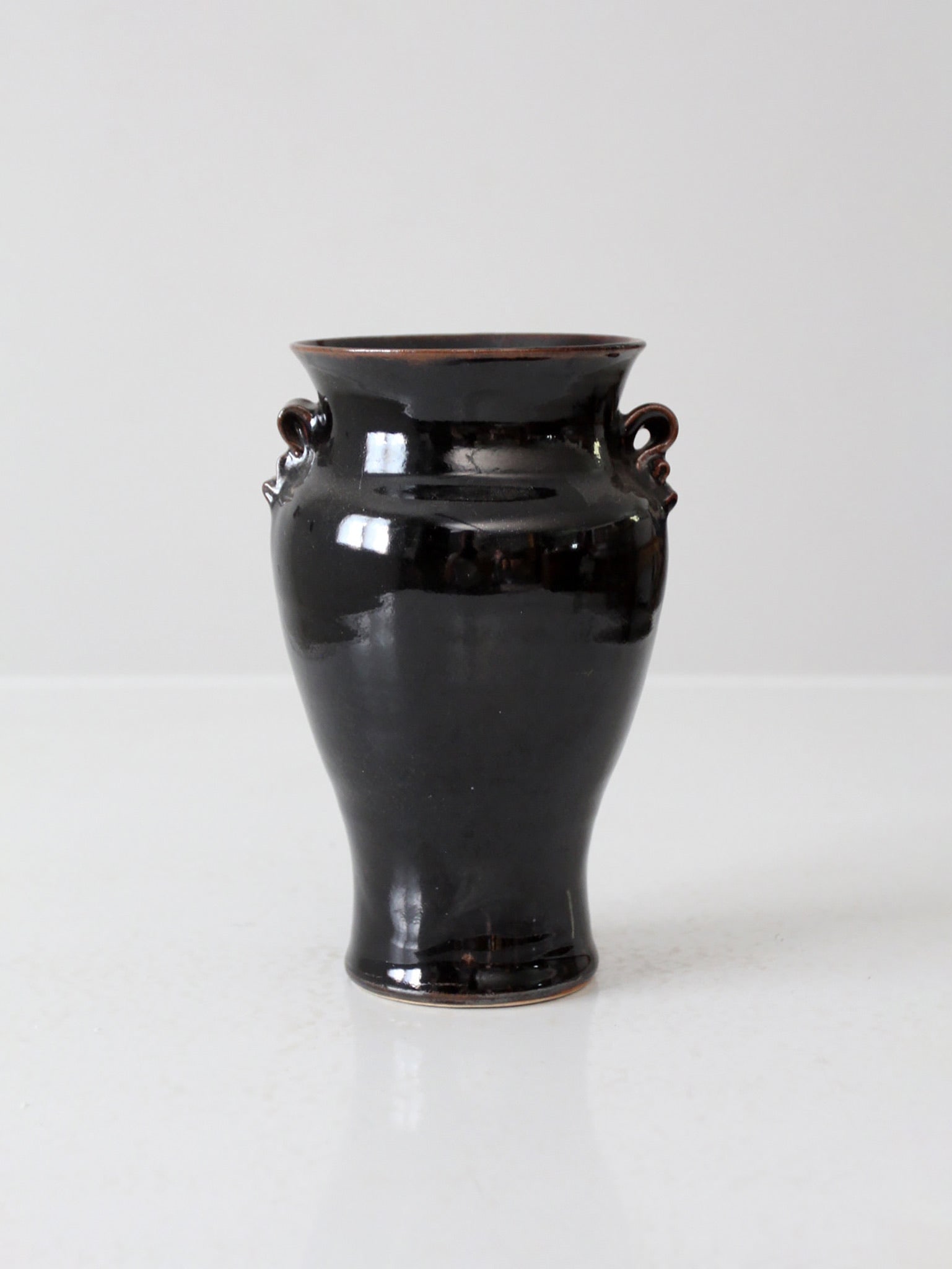 Signed brown glaze studio ceramic vase with loop handles: Signed brown glaze studio ceramic vase with loop handles This vintage studio pottery vase features a rich, deep brown glaze that beautifully captures light, imparting a warm glow. The flared lip and s