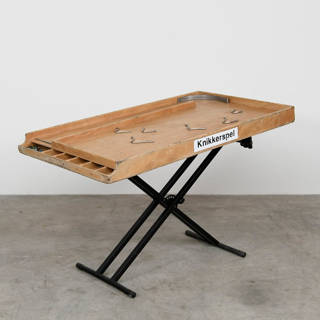 Vintage Traditional Dutch Marble Game Knikkerspel with a Metal Stand: Vintage Knikkerspel, a traditional Dutch marble game table. It features a wooden playing surface with designated scoring pockets and metal pins to obstruct the marbles. The table is supported by colla