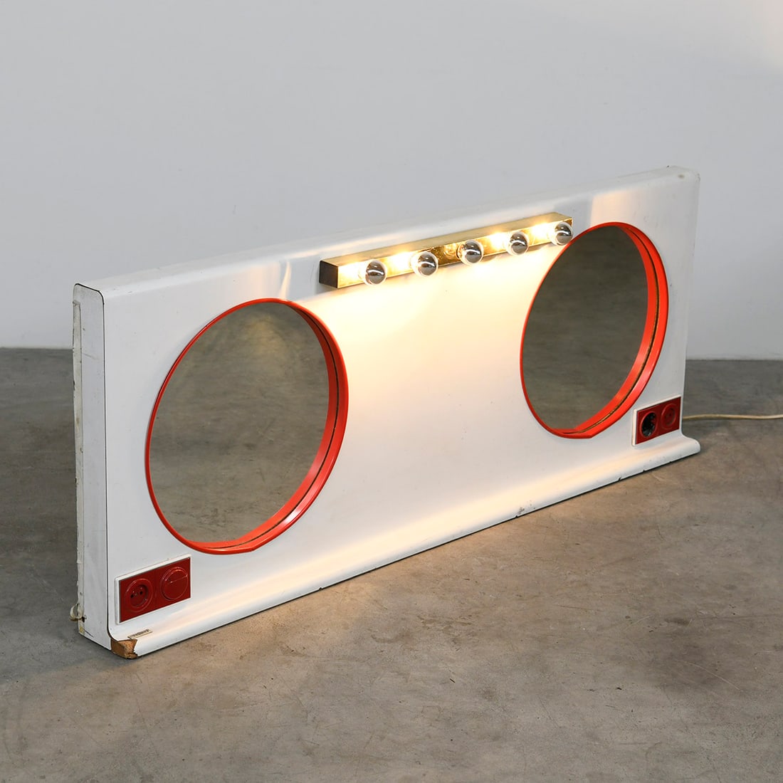 Double Vanity Mirror Featuring Two Round Mirrors, Central Light Bar and Power Outlets: Double vanity mirror unit featuring two round mirrors with red frames mounted on a white panel. The central light bar holds five bulbs, providing bright illumination typical of dressing rooms or backs