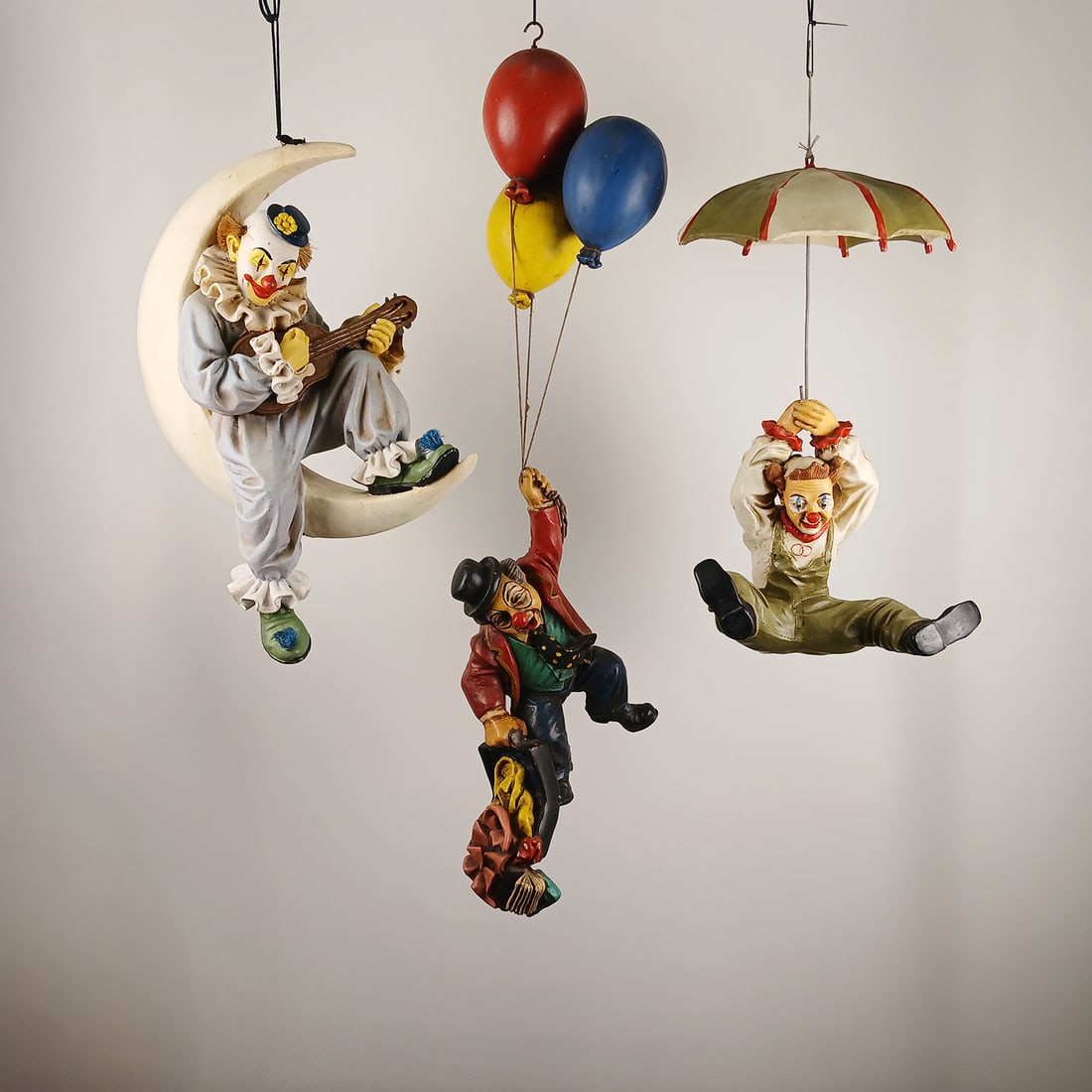 Lot of 3 Hanging Clown Figures: Decorative lot of three medium-sized hanging clown figures. The set includes a clown sitting on a half-moon playing a guitar, a clown hanging from three colorful balloons while holding a suitcase,