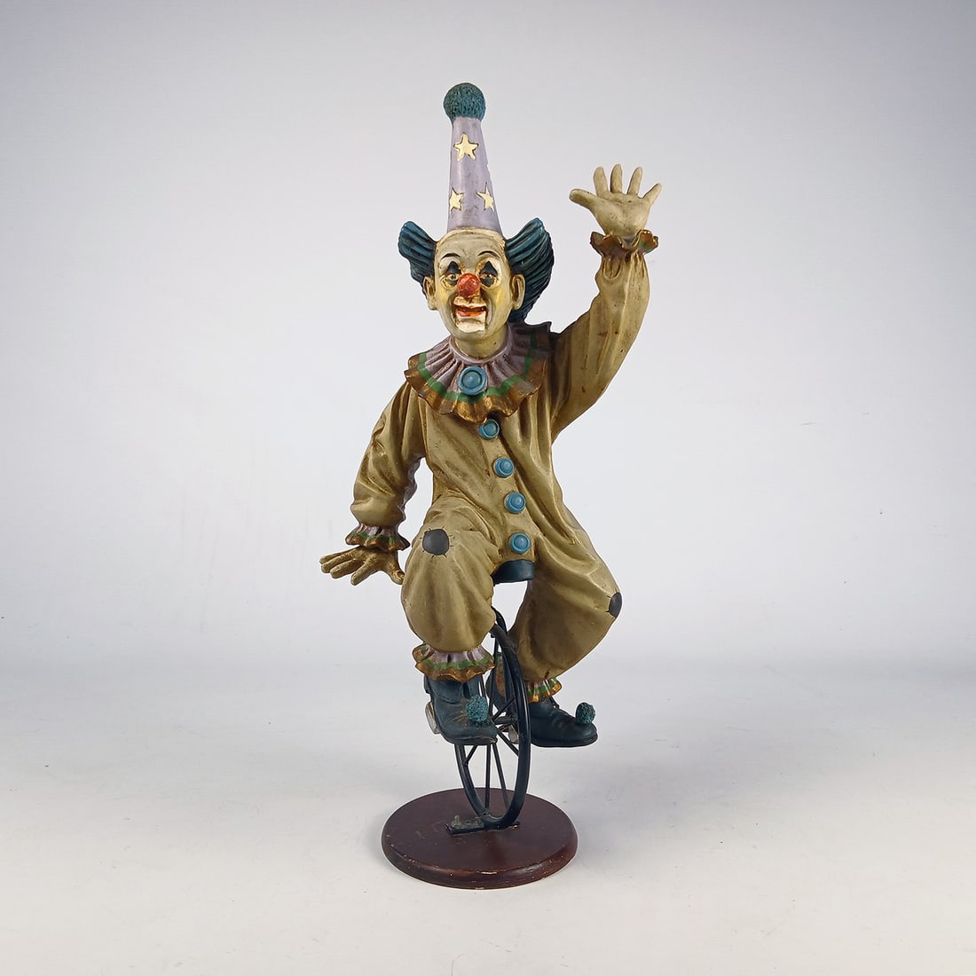 Clown with a pointed hat on a unicycle: Decorative figure depicting a clown balancing on a unicycle while waving. The clown wears a tall pointed hat decorated with stars and a classic circus-style costume, capturing a playful and lively