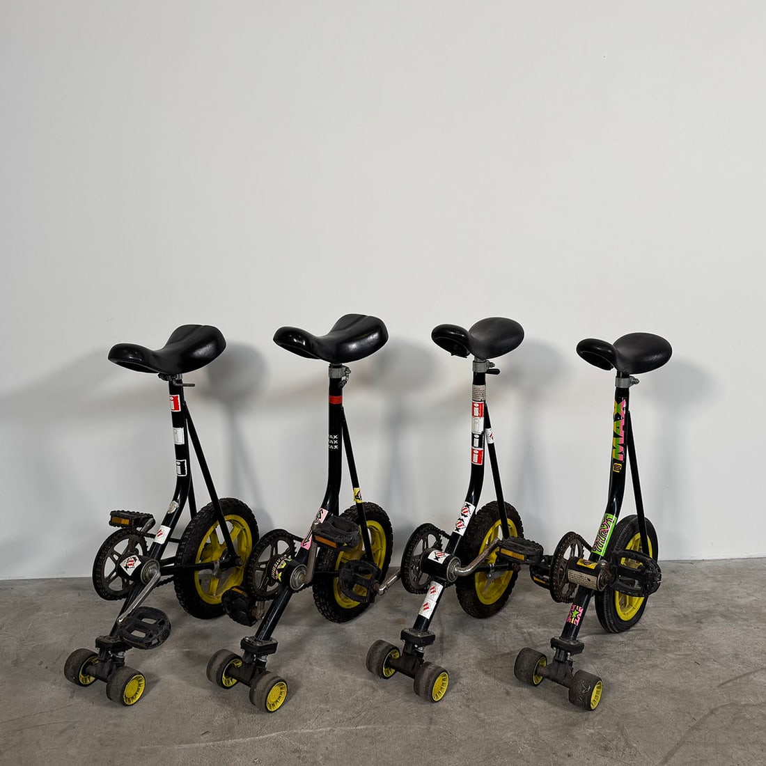 Lot of 4 "Max" Skatebikes: Lot of 4 "Max" Skatebikes, notable for their unique design featuring a unicycle-like frame paired with skateboard-style front steering wheels. Good visual condition, showing some wear. Dimensions (H
