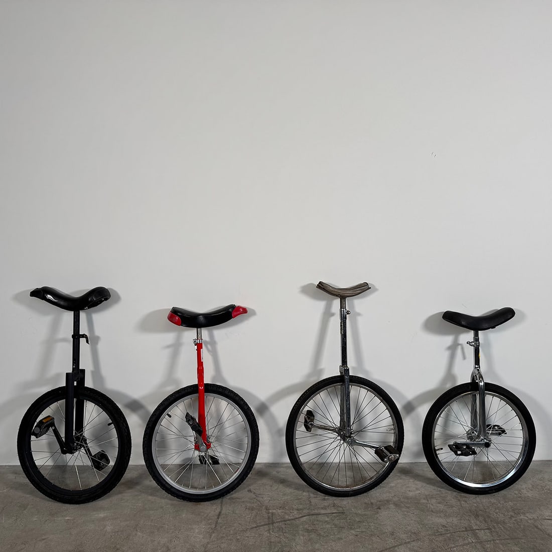Lot of Four Unicycles: Group lot consisting of four unicycles in different colors and designs, each equipped with a saddle seat, pedals, and single wheel construction. These unicycles are typically used for circus training,