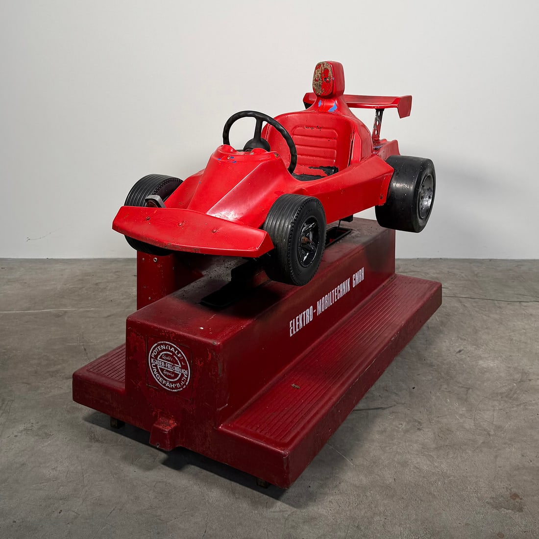 Vintage Red Formula Racer Kiddy Ride (Not Working): Vintage children’s amusement ride designed in the shape of a Formula racing car. The ride features a bright red body with a single molded seat, steering wheel, and racing-style rear wing, mounted on