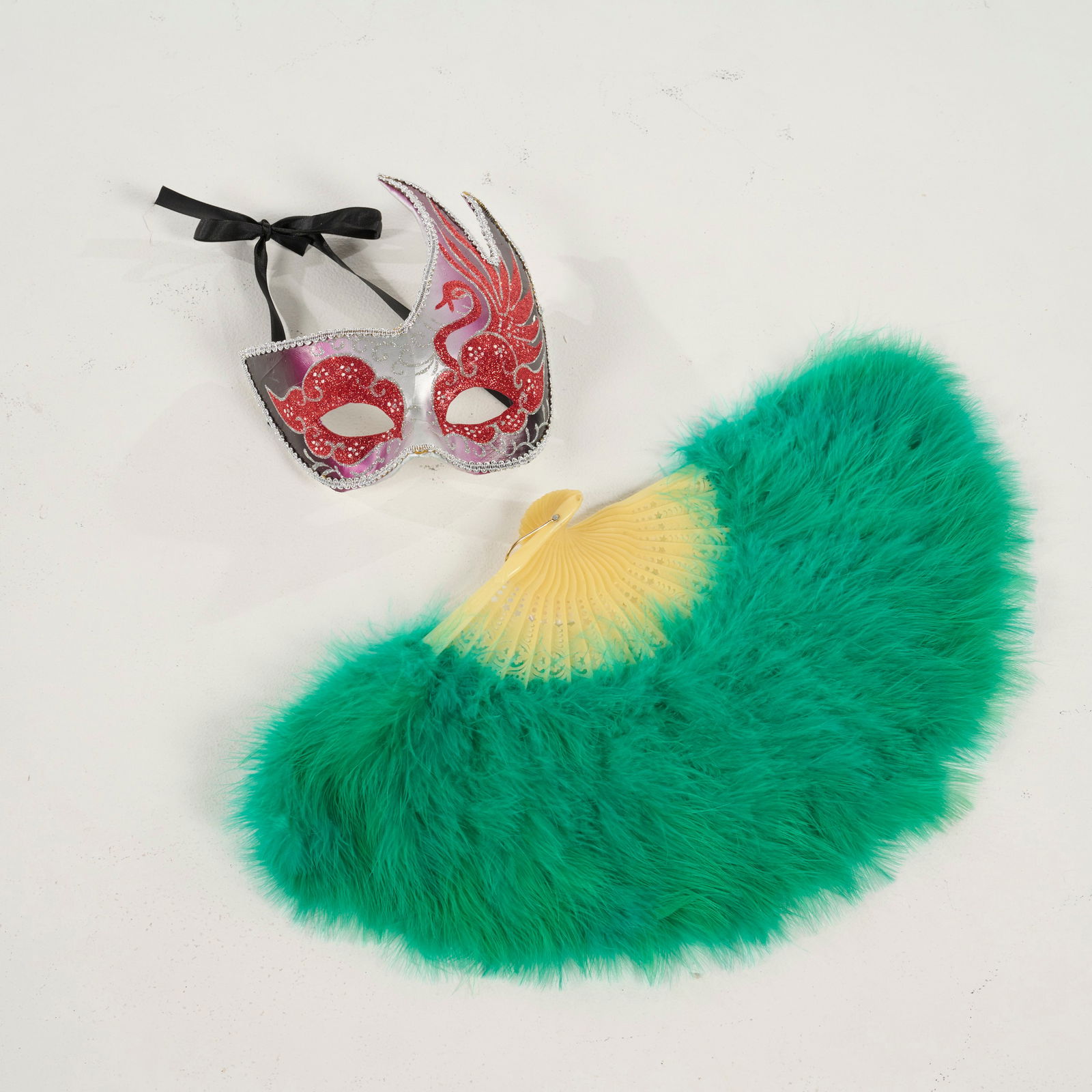 Decorative Mask and Fan Set from the Royce Collection: Shimmering masquerade mask and a vibrant, emerald green feather fan from the Royce Collection. The mask is decorated with red glitter patterns, finished with a silver scalloped trim and black ribbon.