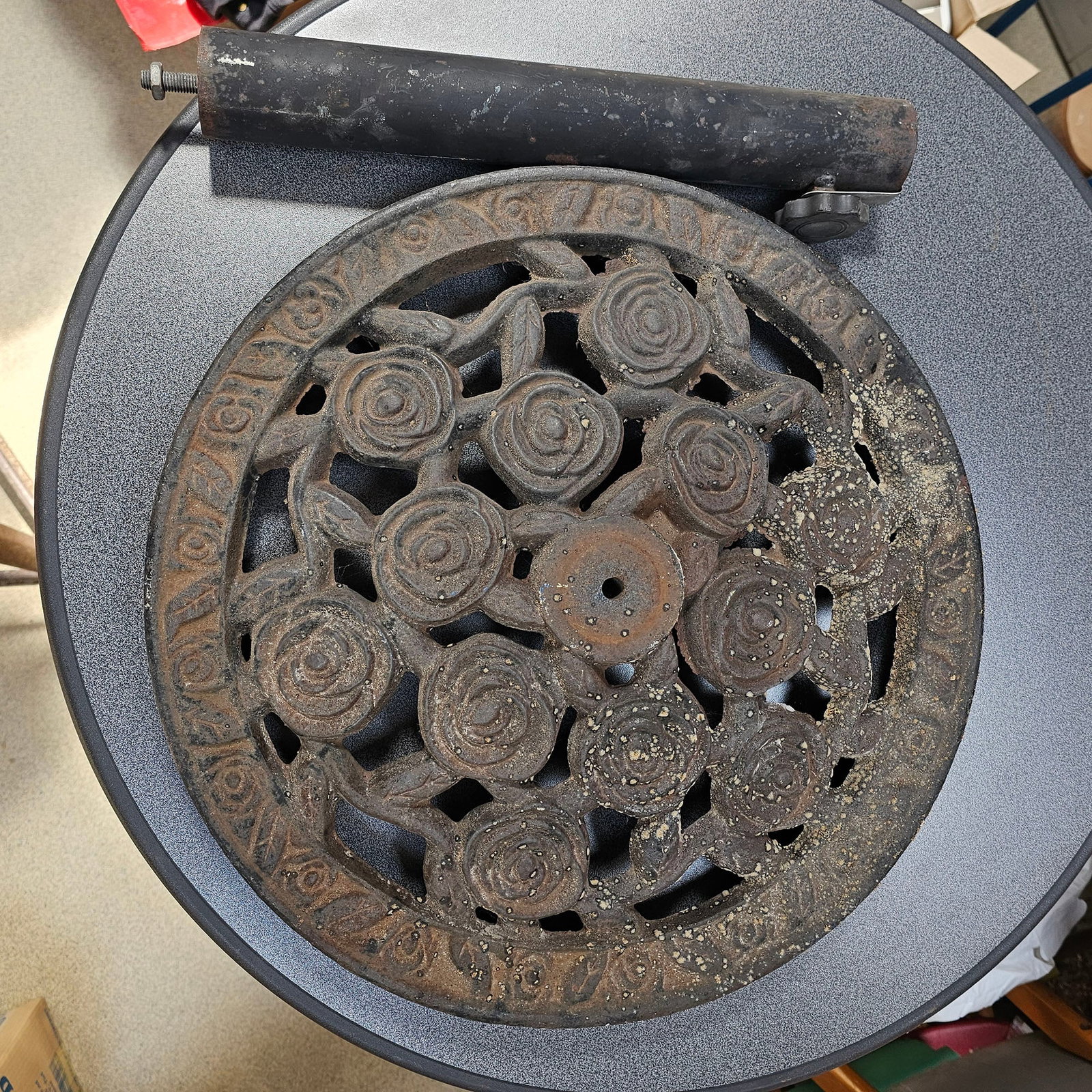 Cast Iron Rose Motif Parasol Base from the G. Perlee Collection: This item is a circular cast iron parasol base featuring an ornate rose and leaf motif throughout its openwork design. It displays significant surface rust and dirt across all surfaces, with some area