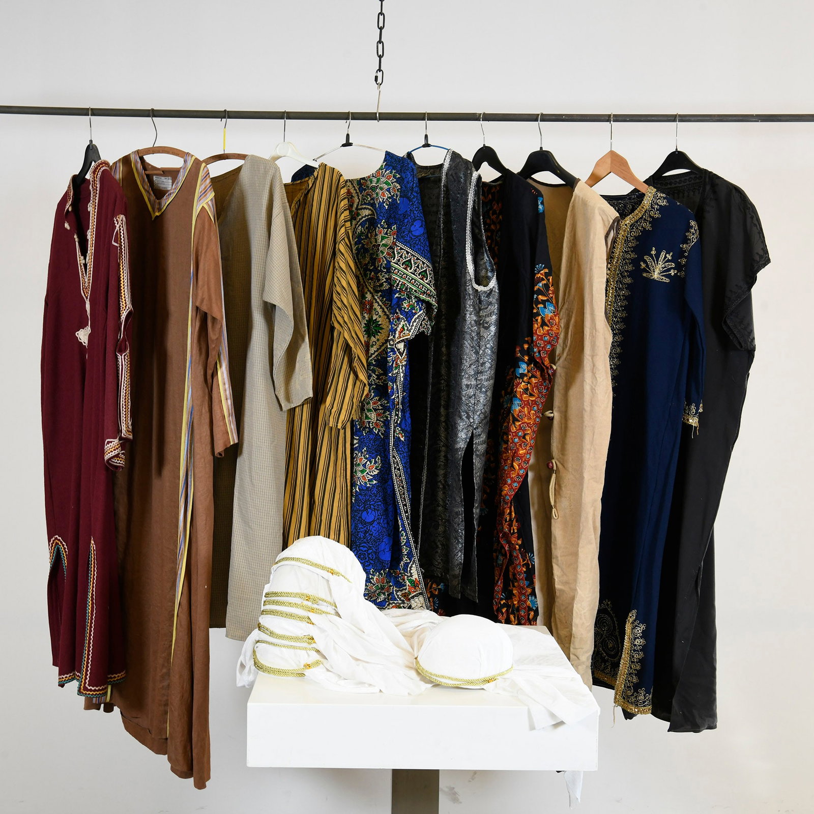 Lot of Assorted Oriental Style Robes & Headpieces: A collection of nine assorted long robes or kaftans and two matching white headpieces. The robes are presented in various colors including maroon, brown, beige, mustard yellow, blue, grey, and black, 