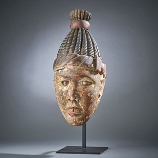 An Nigerian Wood Mask: Southeast Nigeria, the hollowed-out face mask with mouth open showing teeth, drilled eyes and hair, braided coiffure, mounted on a metal stand. Ht. 18, with stand 22 1/4 in. Provenance: Purchased by c