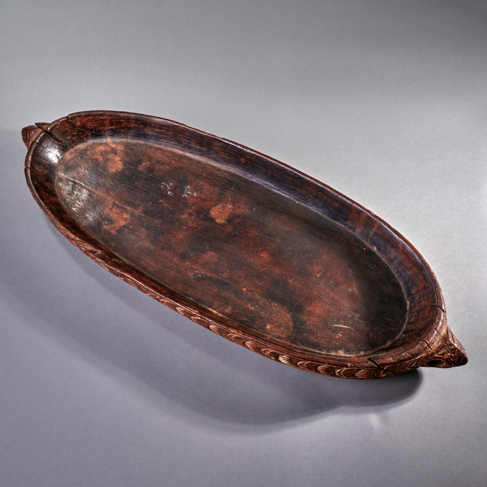 A New Guinea Food platter: Middle Sepik River Province, Papua New Guinea, late 19th Century, wood, decorated around the outer perimeter with scale-like designs, a large lug on each end, smooth, dark patina, minor wood loss. Lg.