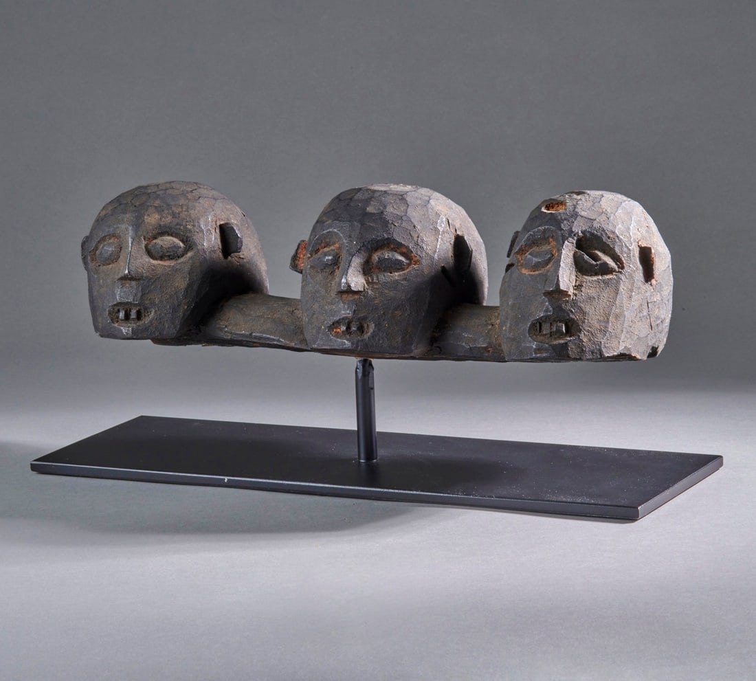 Three Naga wood headhunter skulls: Nagaland, late 19th/early 20th century, three wood skulls carved from one piece of wood, each with mouth open, teeth bared, simple nose and raised oval eyes, dark encrusted patina, mounted on a metal 