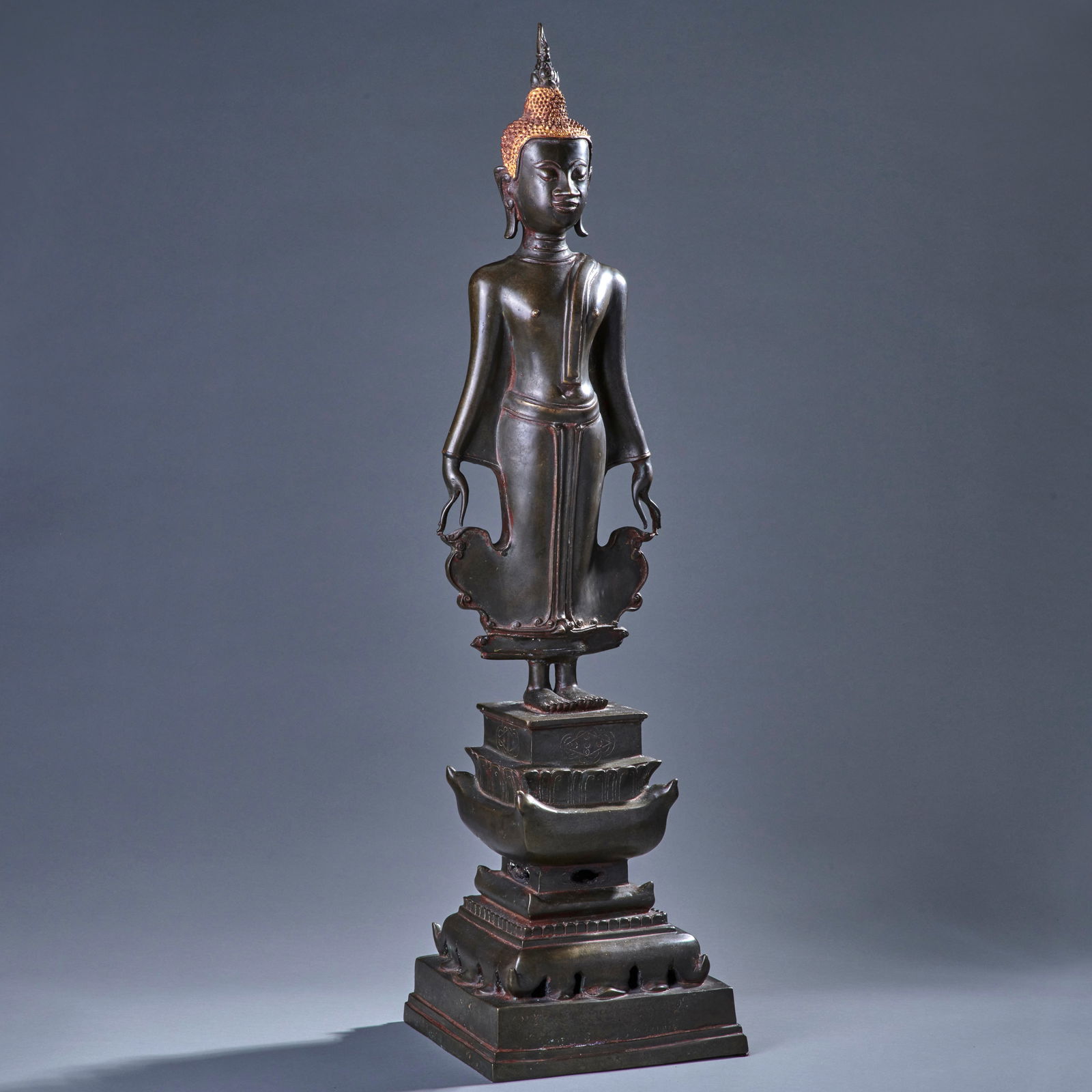 A Large Laotian Standing Buddha: Circa 18th-19th century, standing on an elaborately tiered base, the figure with elongated hands, is in the distinctive ‘calling for rain’ mudra which is unique to Laotian works. Small repair to f