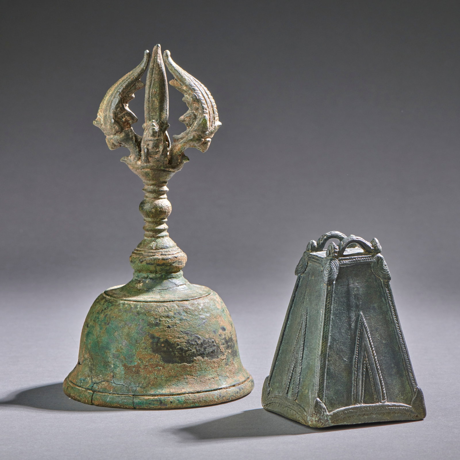 Two Khmer Bronze Bells: Circa 12-13th century, a Bayon style handbell with 2 opposing seated figures on vajra finial. The other, of trapezoidal form with delicate linear and spiral decorations. Both fine examples. Handbell h