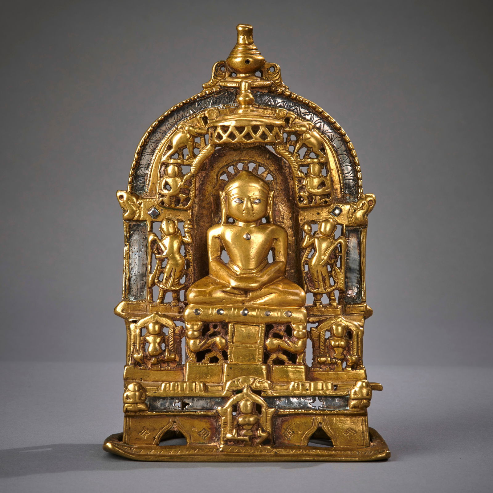 A large Indian Brass Jain Shrine: Gujarat, India, circa early 16th century (dated 1517), of typical form, the seated Jina surrounded by openwork attendant figures, deities and elephants, with copper and silver inlaid features, embelli