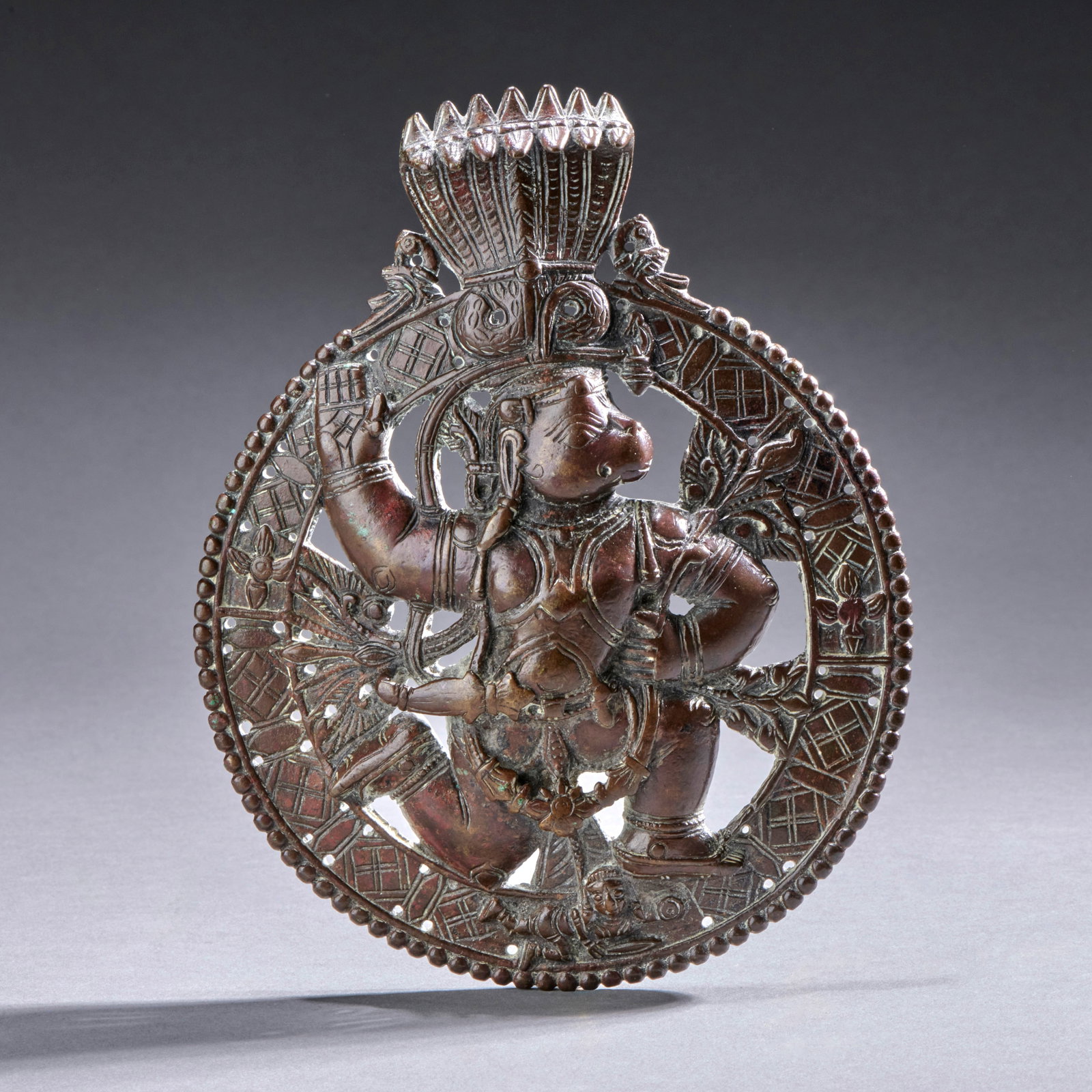 A large Indian Bronze Hanuman Plaque: India, circa early 19th century, the openwork roundel of typical form, with an animated Hanuman trampling the goddess Lanka while grasping an herb bundle, his head protected by a 7-headed Naga framed 