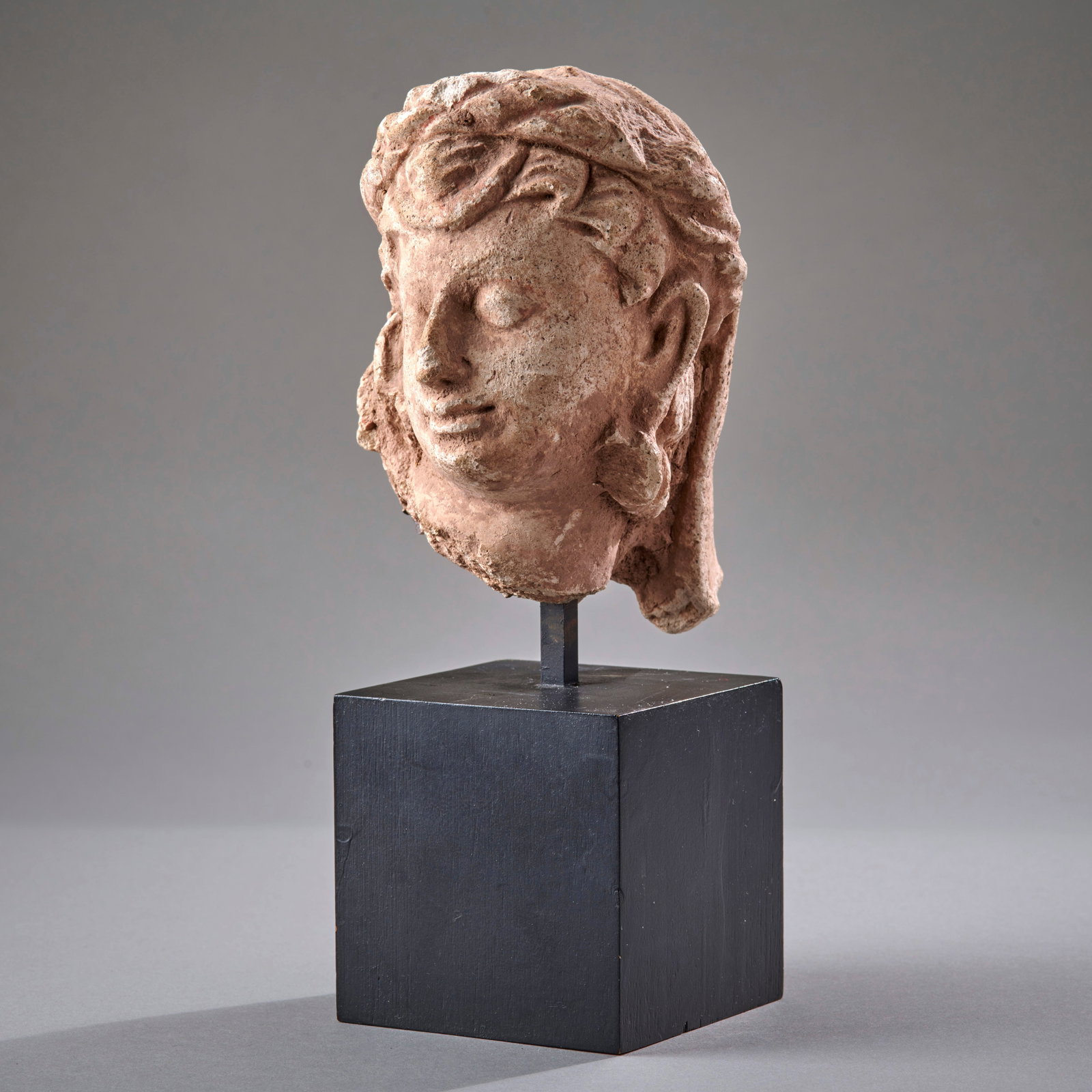 A Gandharan Stucco Head: Circa 2nd to 4th century A.D., a sensitive portrait of a youthful deity or adorant, with flowing hair headdress. Mounted on a custom base. In good condition, with wear. Ht. 5 3/16 in. ht. with base 8 