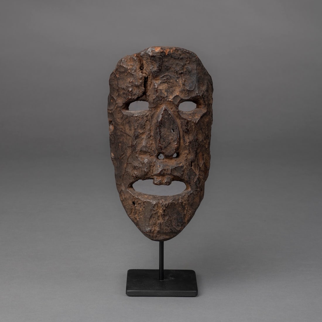 A Nepal mask: The small mask with single pierced hole on either side, with curved face, large open mouth with two teeth, flat nose and cutout eyes, black, encrusted patina, on a metal stand. Ht. 7 3/4, wd. 4 1/4 in