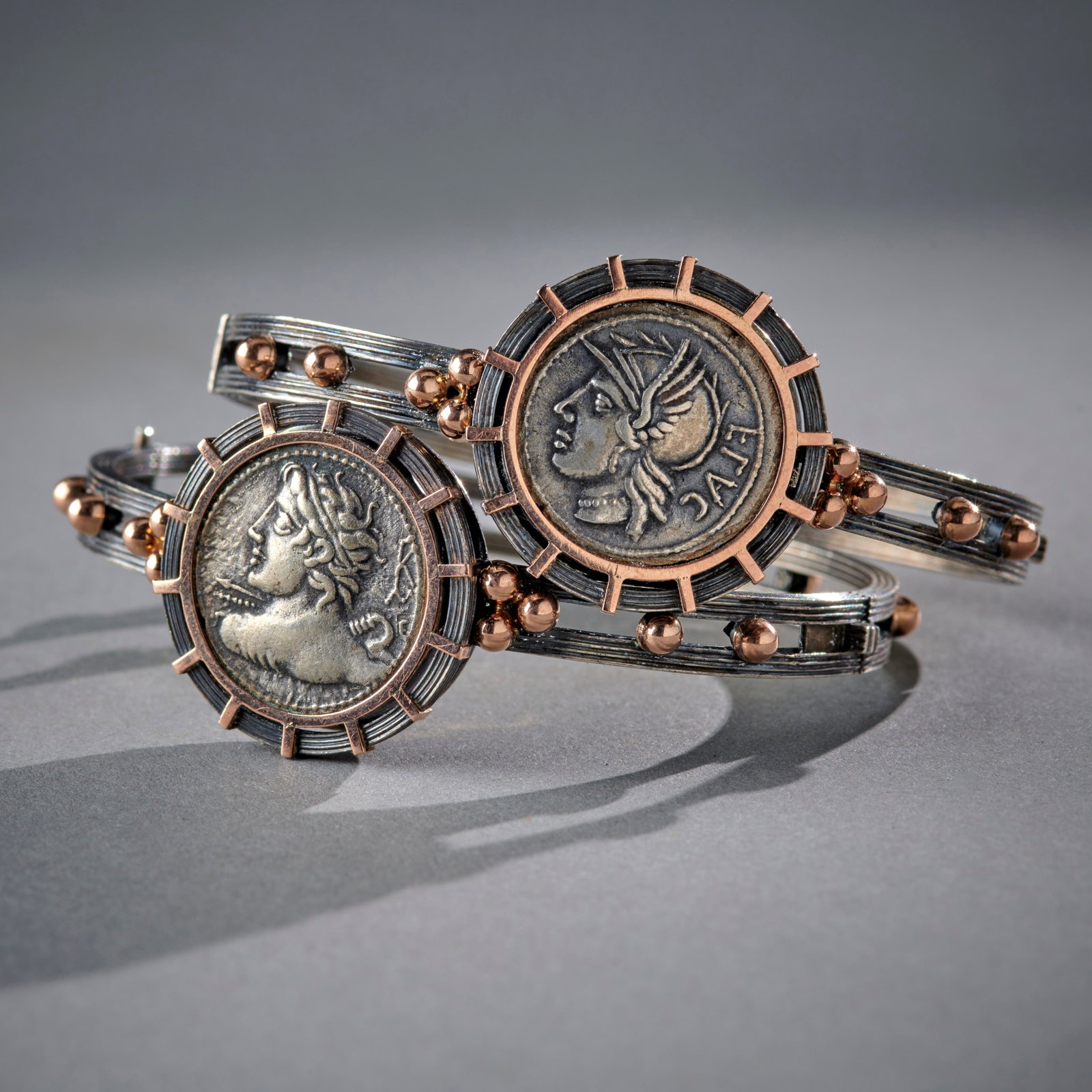 A Pair of Silver Etruscan Revival Bracelets: Late 19th century, European, the hinged silver bands accented with gold spheres, each mounted with an ancient silver coin, surrounded by a spoked circular gold mount. In good condition. Dia. 2 7/16, i