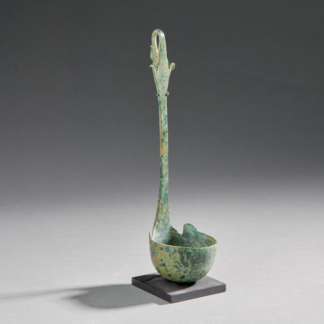 A bronze Hellenistic ladle: Asia Minor, circa 2nd/1st century B.C., raised from sheet bronze, the ladle with a deep bowl, spurs and a duck head terminal on the handle., with dark glossy patina, custom stand. Intact. Lg. 7 in. wi