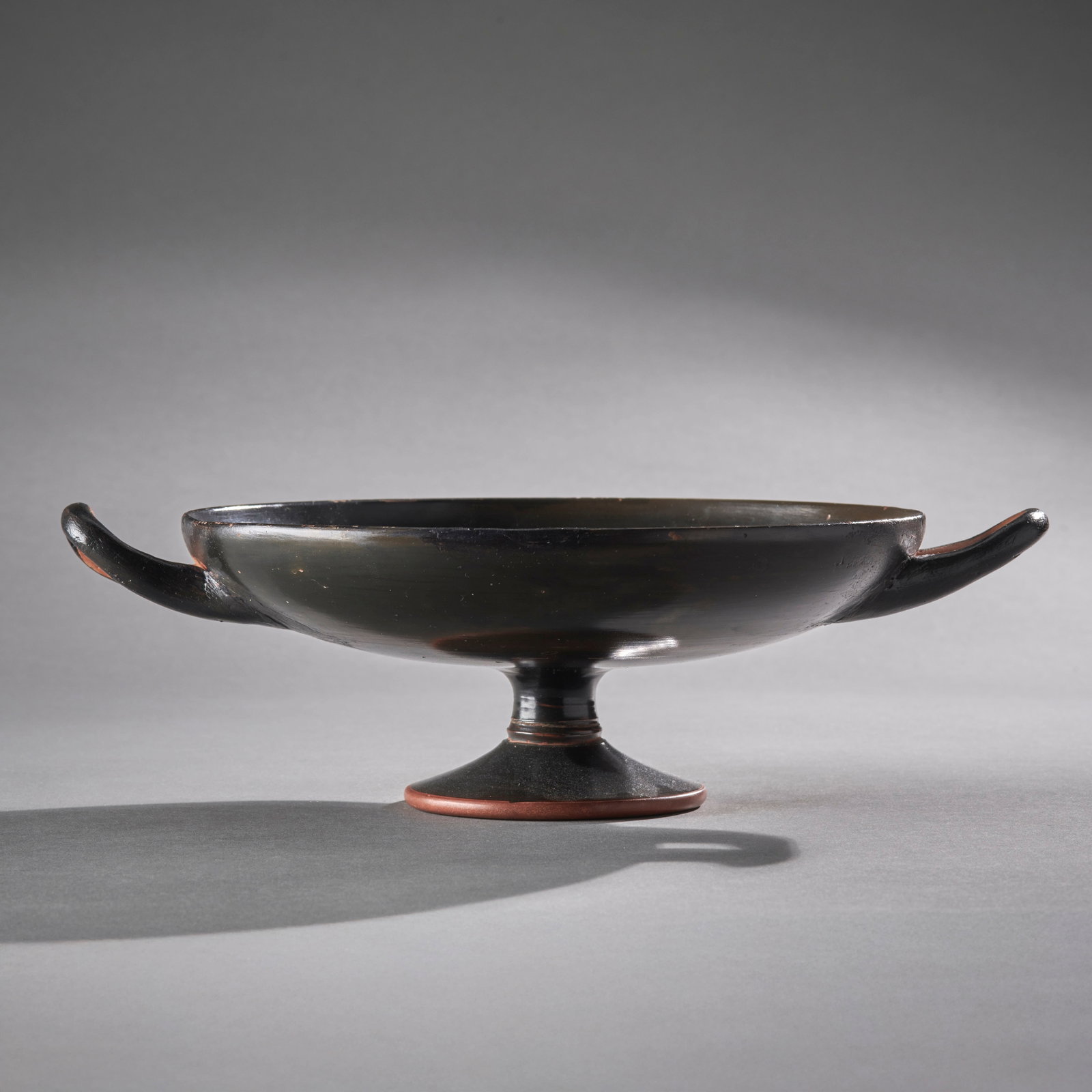 A Greek Terracotta Kylix: Likely of Attic origin, circa 5th/4th century B.C., an elegant bowl sitting on short stemmed foot, with twin looped handles and a vibrant dark metallic glaze. Repair and possible restoration to one ha