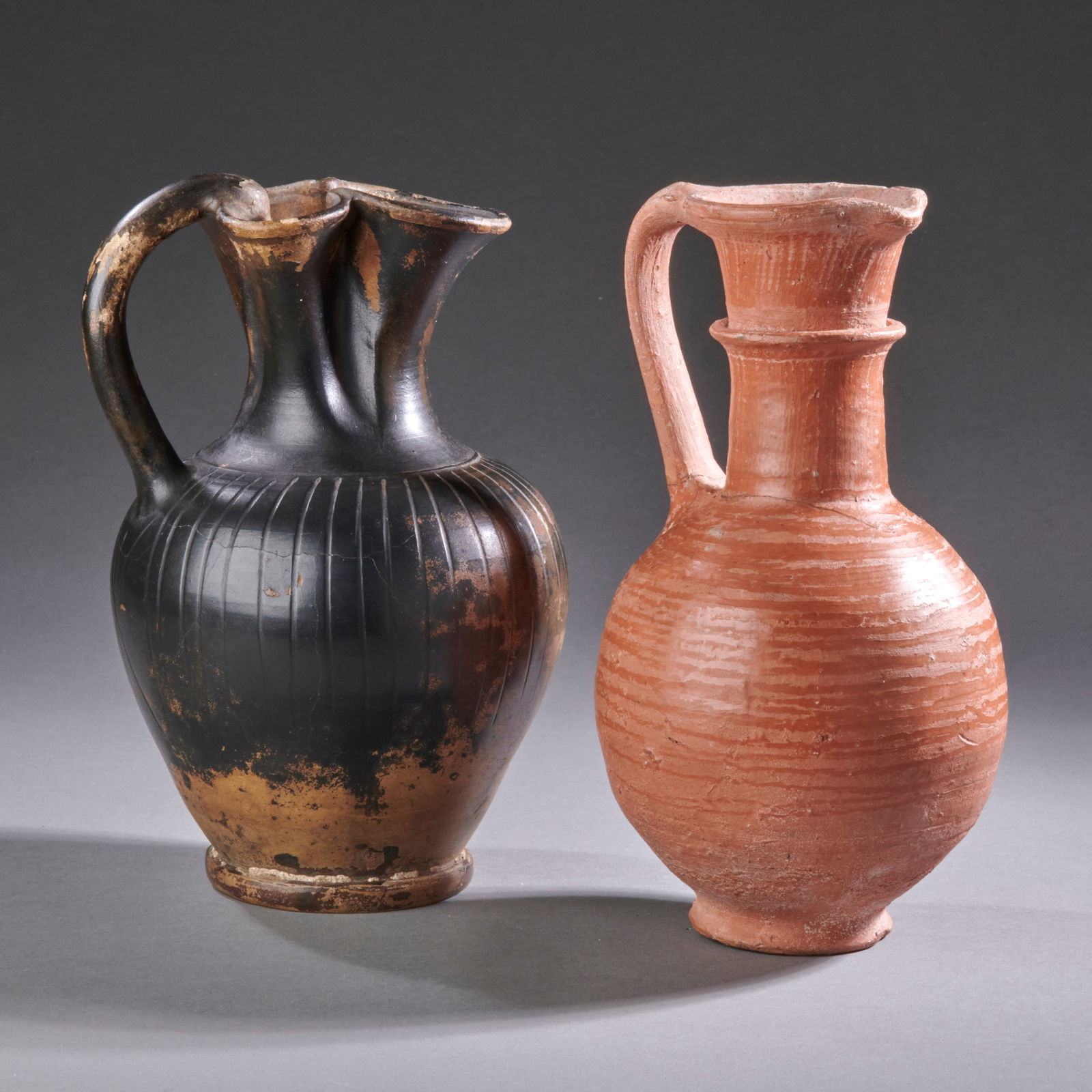 Two Greco-Roman Ewers: Magna-Graecia, circa 4th century B.C.-1st century A.D., a Greek black glaze ribbed oinochoe and a burnished red ware ewer. Oinochoe with stable crack, the ewer repaired from two pieces. Ht. 8 in. each