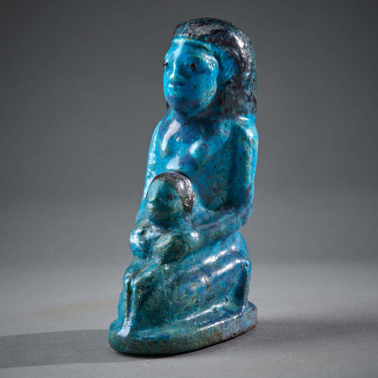 A Large Egyptian Faience Isis Horus Group: Roman period, circa 100 B.C. -100 A.D., the bi-chrome faience statuette of a woman kneeling on an oval, flat cushion and holding a child, reminiscent of New Kingdom milk vessels used in the healing of