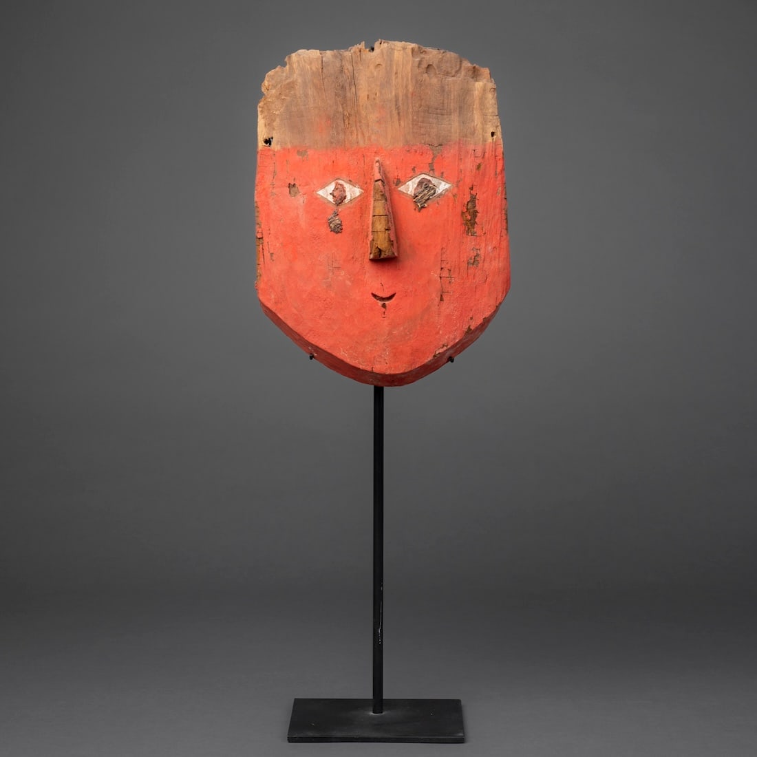 A Chancay polychrome wood mask: Chancay, circa 1000-1250 A.D., the wood mask, with red pigment on the face, the diamond-shaped eyes with cloth inlay, and simple raised nose, mounted on a metal stand. Ht. 10, wd. 7 1/2 in.Provenance: