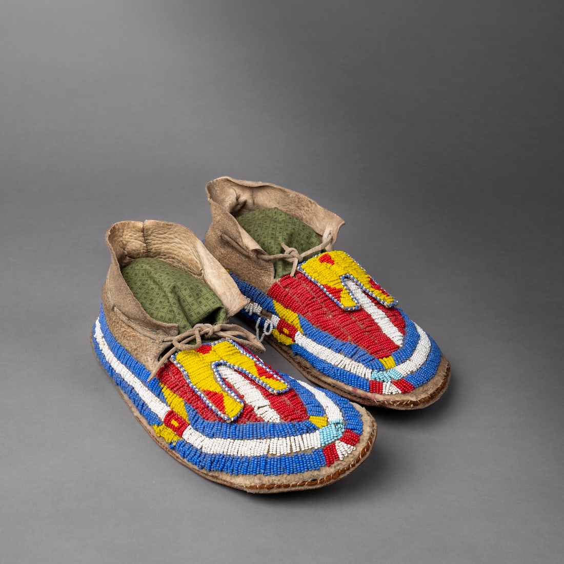 A pair of fully beaded Plains moccasins: Lakota, (Sioux), post 1900, the moccasins with hard leather soles with fully beaded uppers and bifurcated tongues in multi colors, with red cloth at the sole seams, (minor bead loss and some loose bea