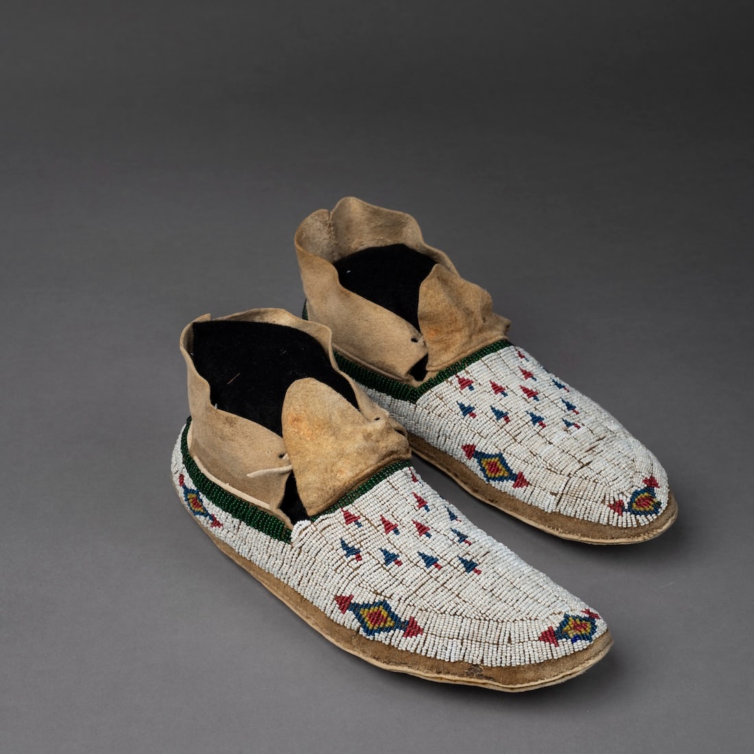 A pair of Northern Plains beaded hide moccasins: Arapaho or Cheyenne, fourth quarter 19th century, the rawhide soles with fully beaded vamps, with arrow point motifs in red and blue on a white bead ground. Lg. 10 3/8 in.