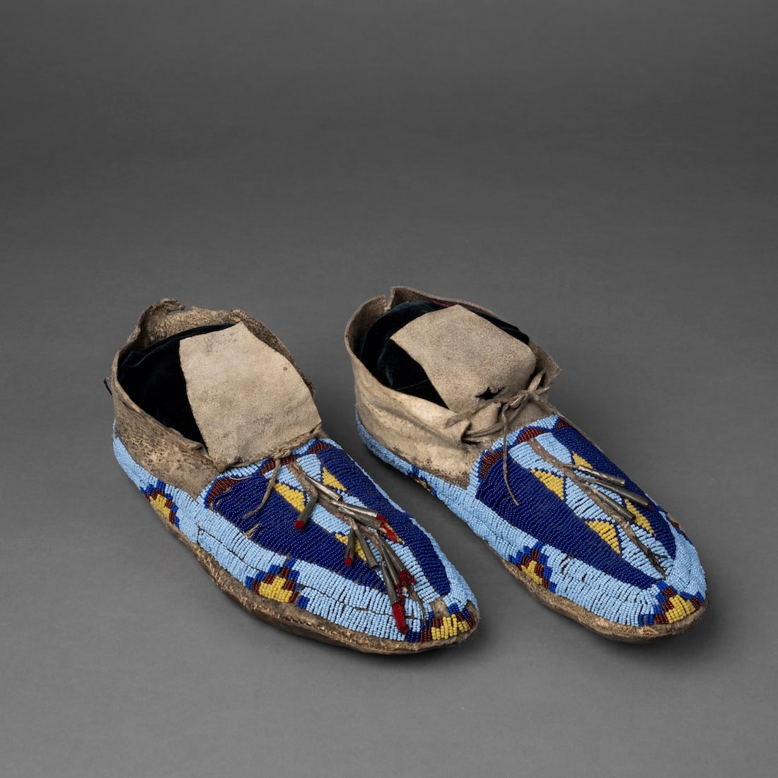 A pair of Plains beaded hide moccasins: Fourth quarter 19th century, decorated on the vamps with multicolored geometric designs on a blue ground, with tin cons in a central strip, with square-cut hide tongues, (bead loss and tin cone loss i
