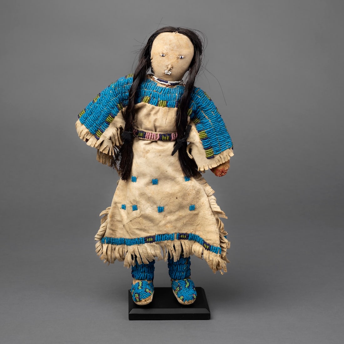 A Plains beaded hide doll: Lakota, (Sioux), Fourth quarter 19th century, wearing beaded hightop moccasins, with long fringed dress with beaded band at the bottom and decorated with small beaded squares, with beaded belt, with a