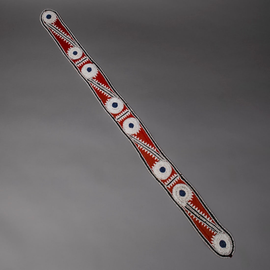 A Choctaw beaded sash: Third quarter 19th century, backed with red trade cloth, decorated on one side in four panels of repeated medallions joined by serrated bands, in white and blue/green beads. Lg. 45, wd. 3 1/4 in. Prov