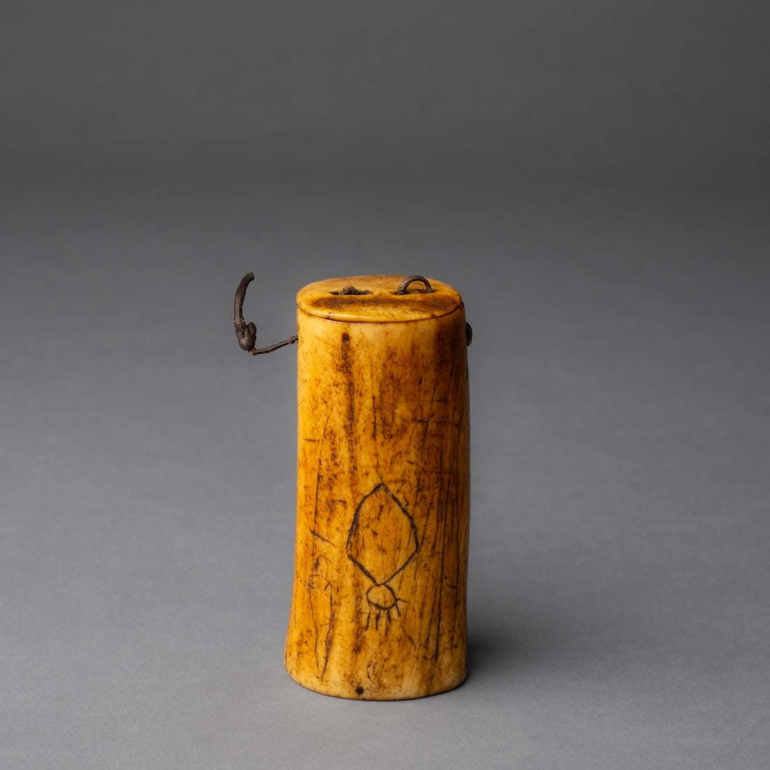 An Eskimo man’s bone container: Alaska, 19th century, the large bone container with a wood base, and bone lid attached with hide, decorated with simple figures and arrow-like design, smooth, dark patina. Ht. 4 1/4, wd. 2 in. Provena